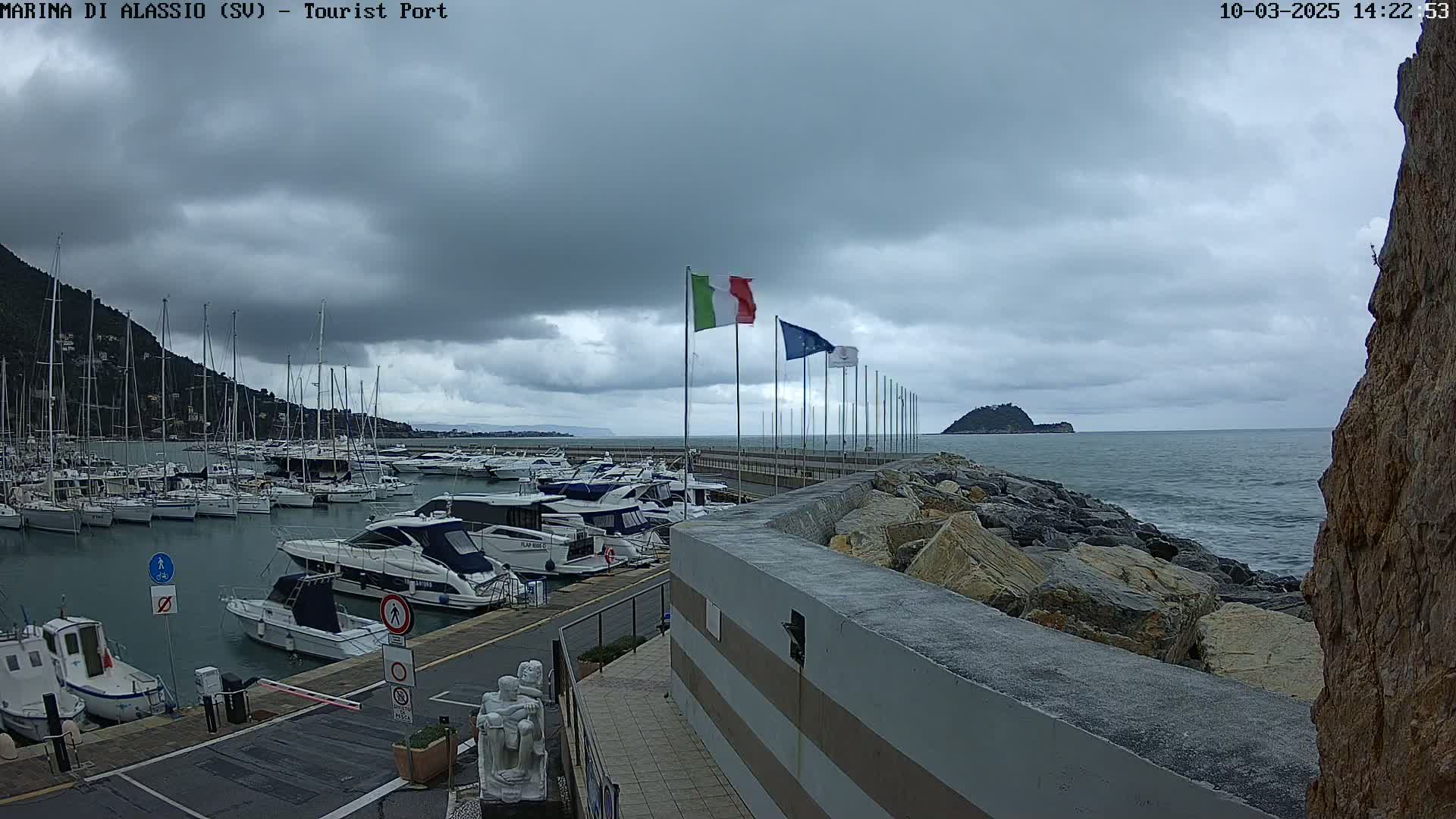 A marina filled with boats sits under a cloudy sky next to a rocky shoreline and a small island in the distance.
