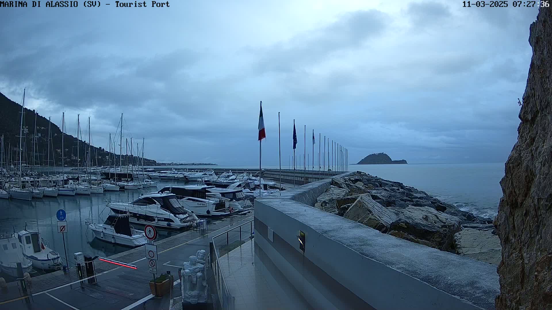 A marina filled with boats is situated next to a rocky shoreline under a cloudy sky.