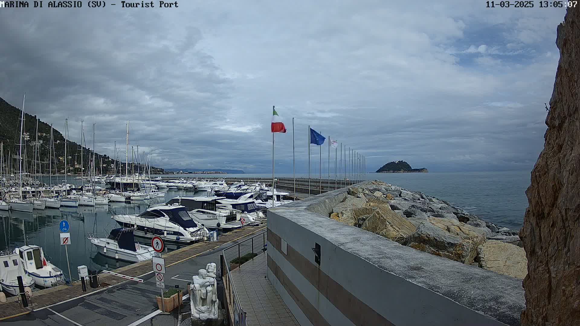 A harbor filled with many boats is seen under a cloudy sky, with a rocky shoreline and calm water extending to a small island in the distance.