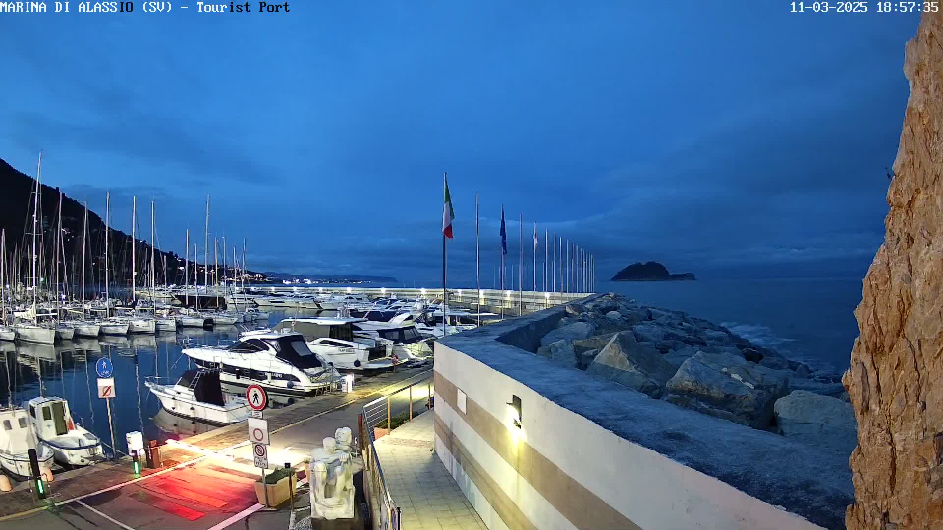 A marina filled with yachts and sailboats sits under a twilight sky, with a rocky seawall and calm ocean visible in the background.