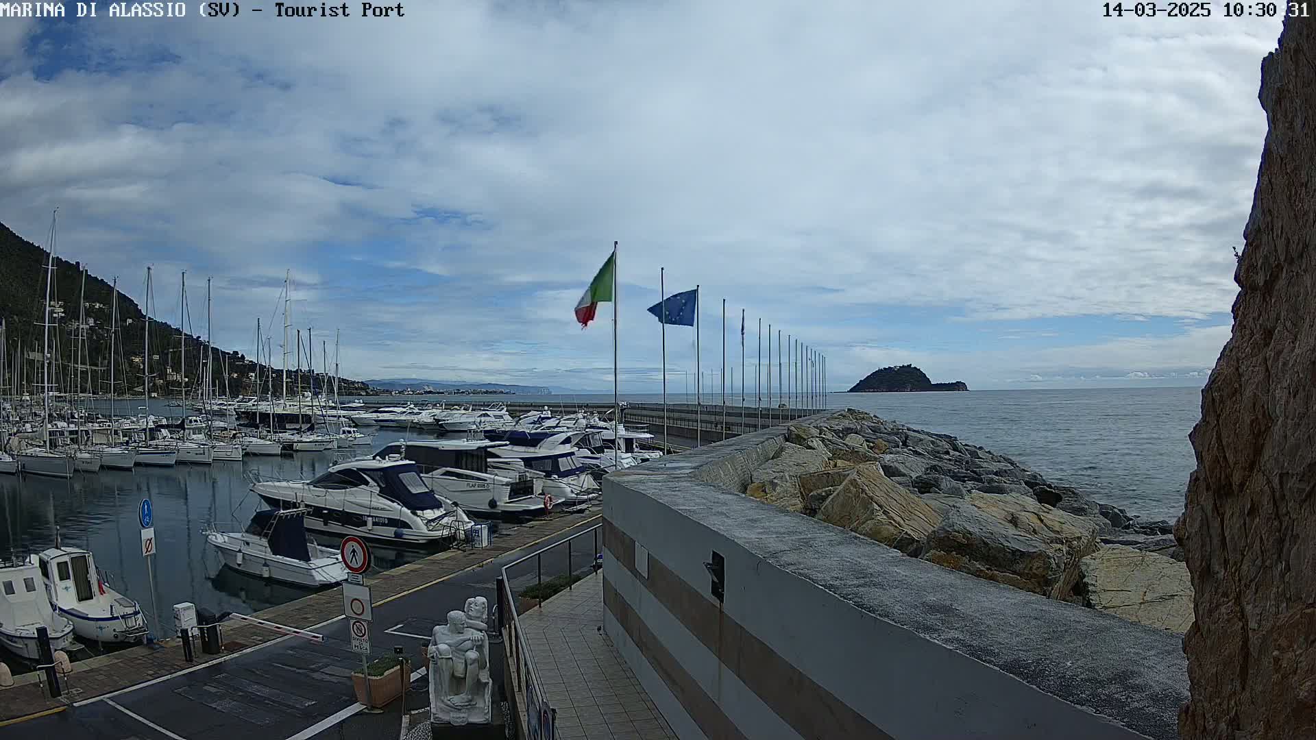 A marina filled with boats is situated next to a rocky shoreline under a partly cloudy sky.