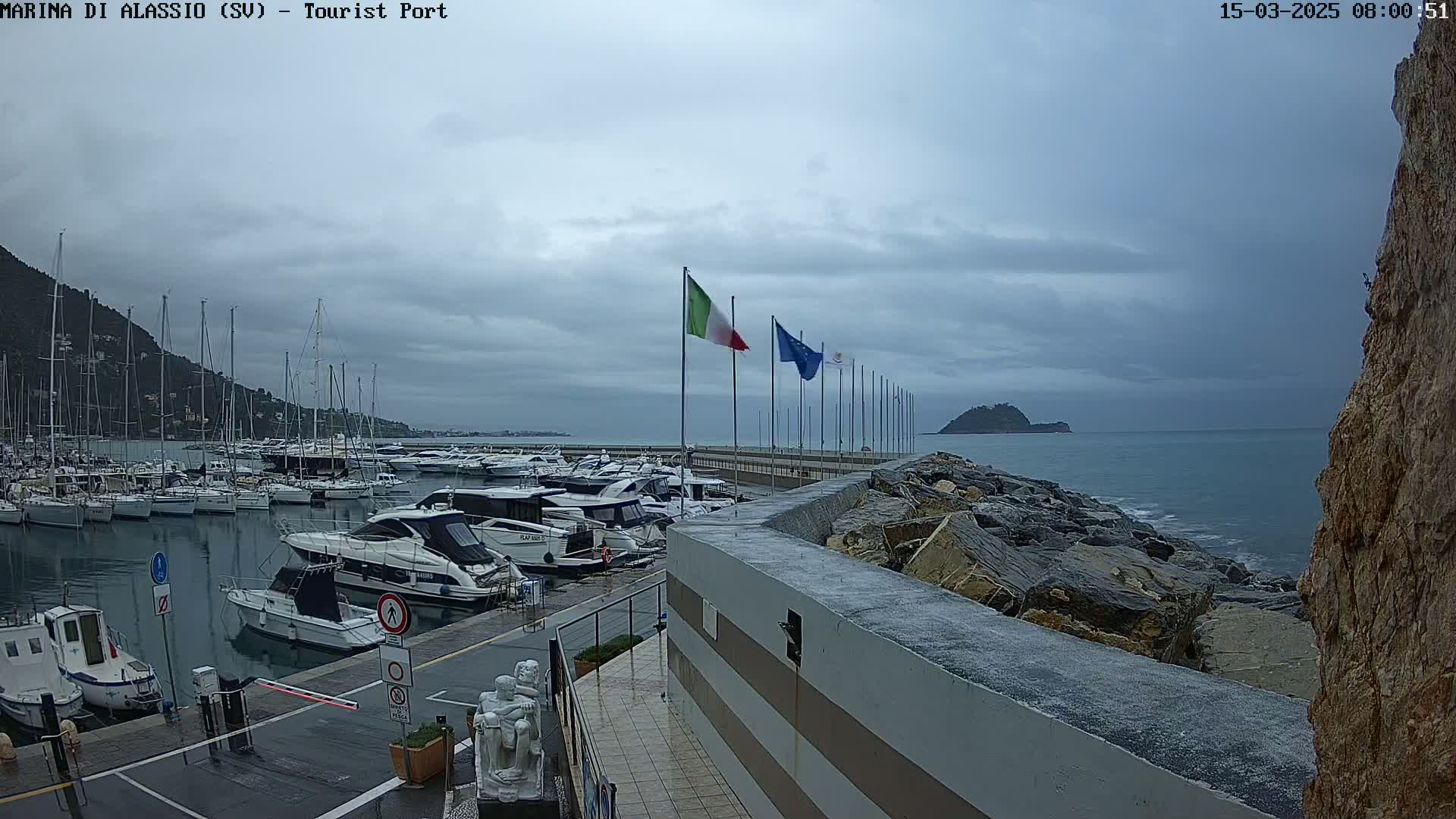 A marina filled with boats is seen under an overcast sky, with a rocky shoreline and calm sea in the background.