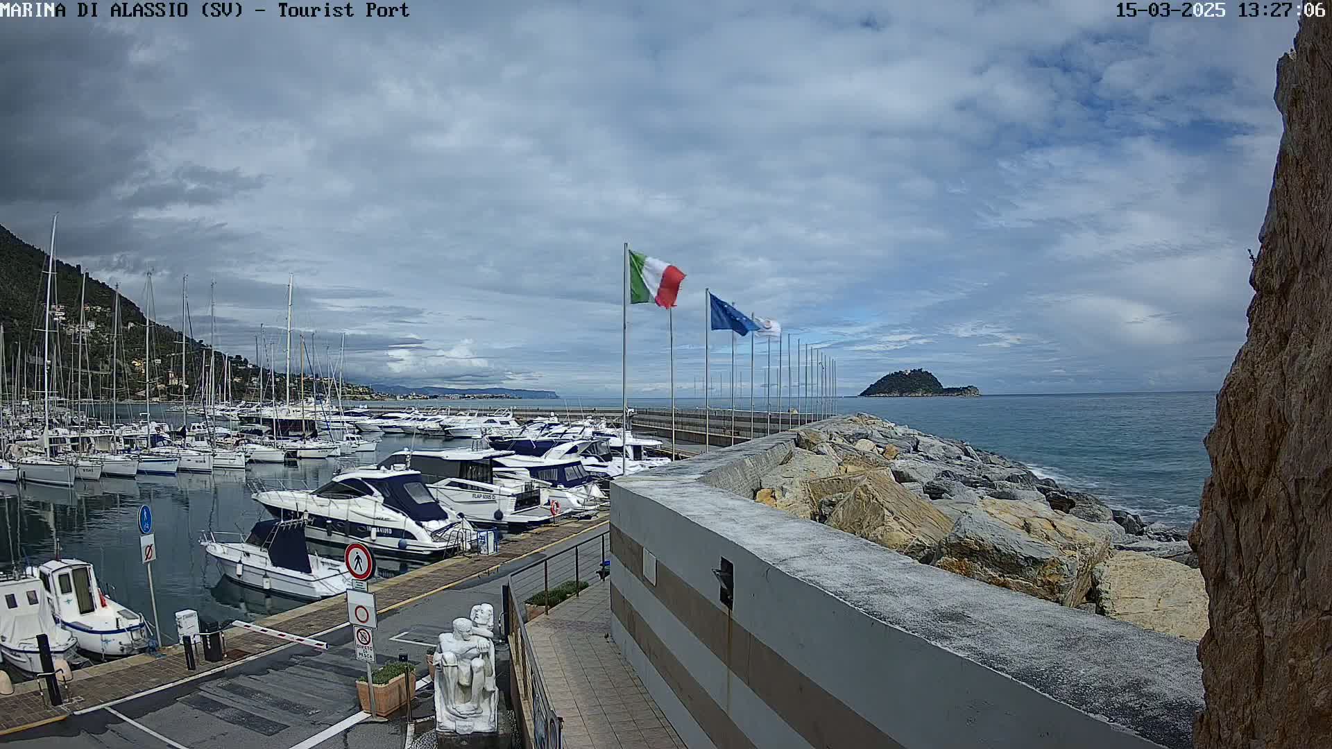 A harbor filled with boats is situated on a partly cloudy day, with a rocky shoreline and an island visible in the distance.