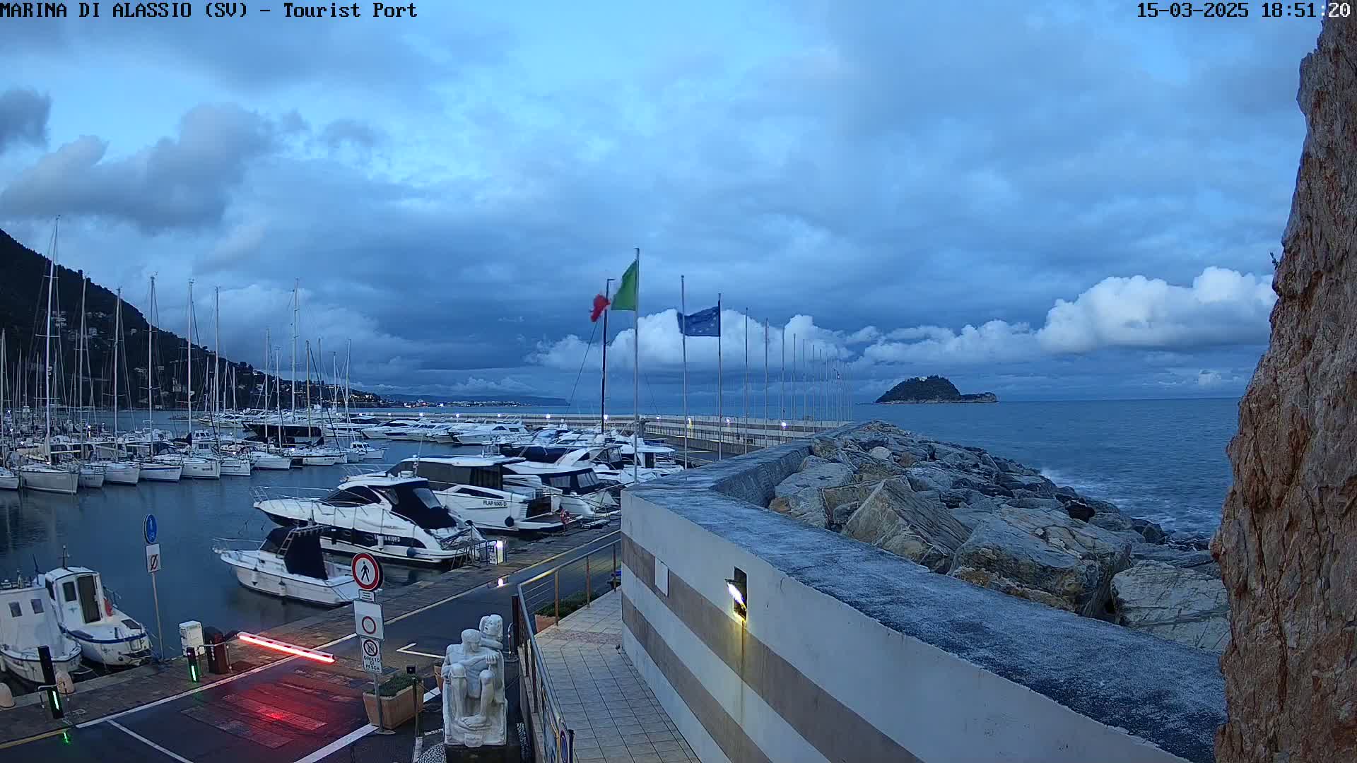 A harbor filled with yachts and boats under a cloudy, twilight sky, with a rocky breakwater separating the marina from the sea.