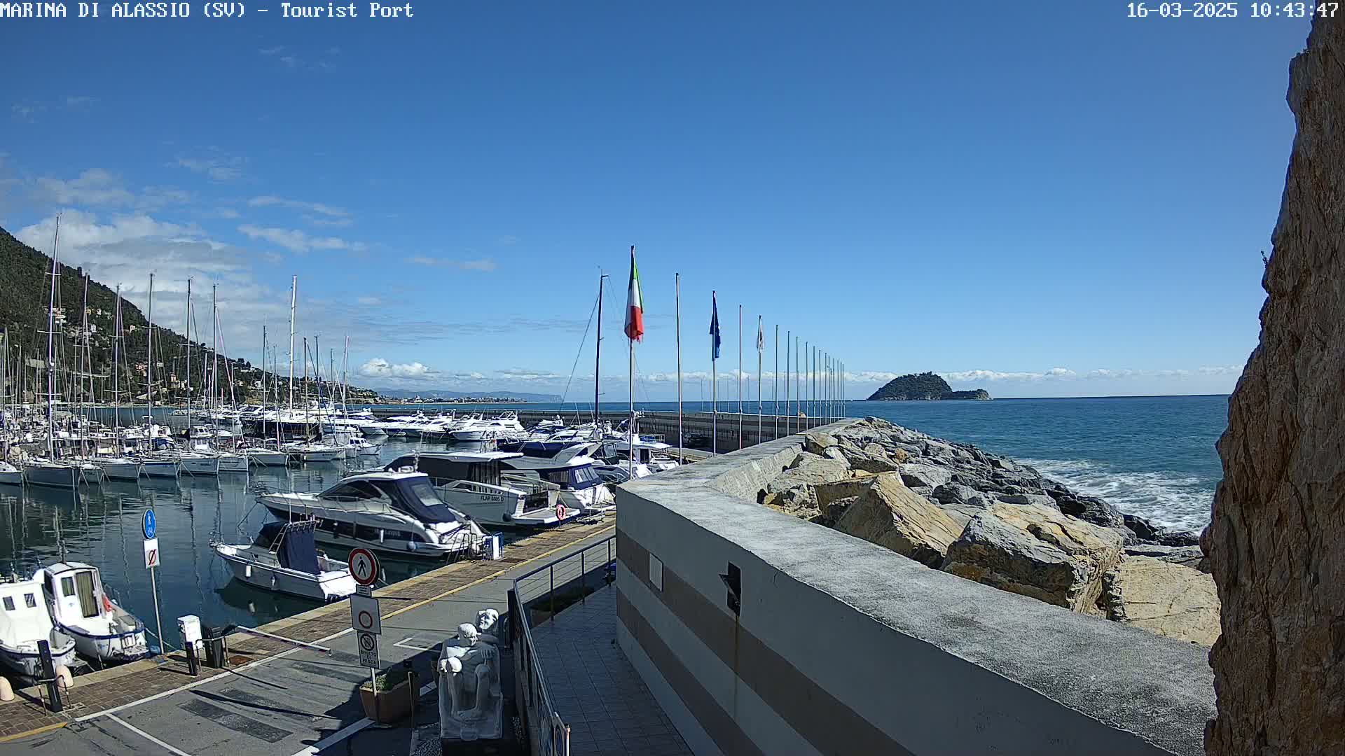 A marina filled with boats is situated next to a rocky shoreline under a clear blue sky.