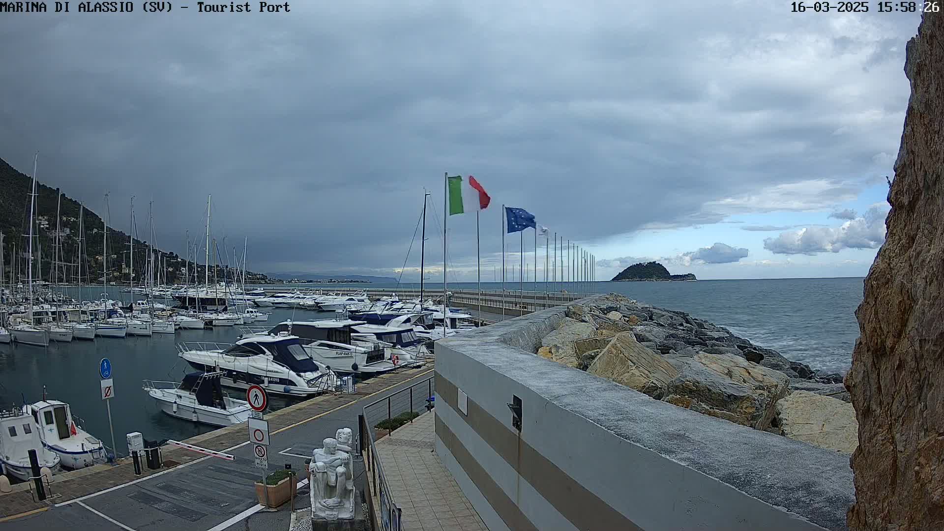 A marina filled with sailboats and motorboats is shown under a cloudy sky, with a rocky shoreline and a small island visible in the distance.