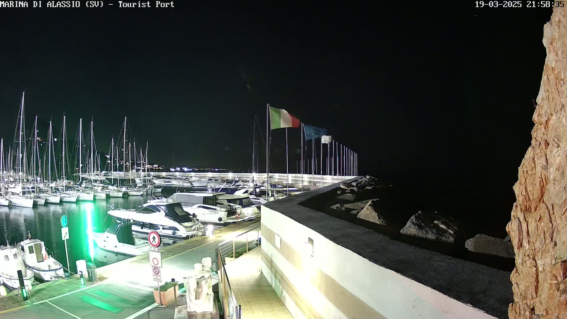 At night, a marina filled with docked sailboats and motorboats is shown, with Italian flags waving gently in the dark sky.