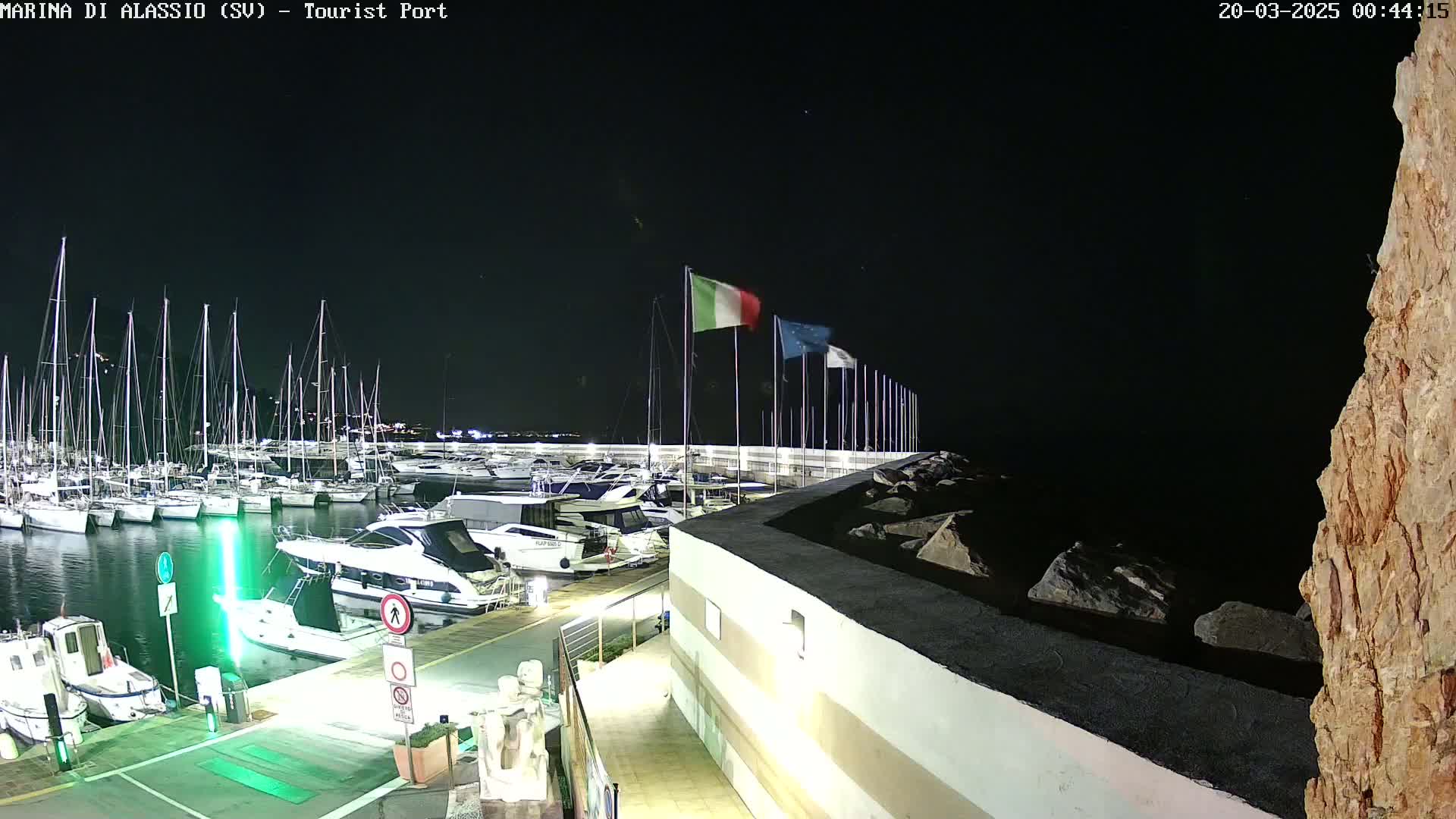 A marina at night, filled with docked boats under a clear, dark sky, with Italian and European flags waving in the breeze.