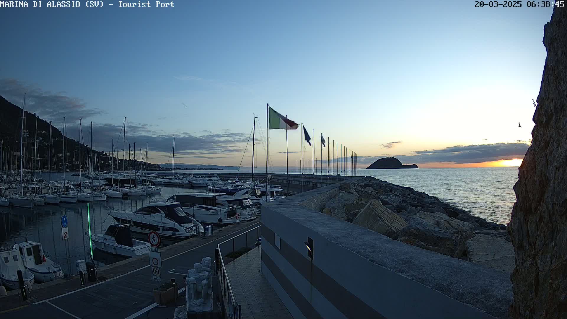 A marina filled with sailboats and motorboats at sunrise under a mostly clear sky, with a rocky shoreline and a small island visible in the distance.