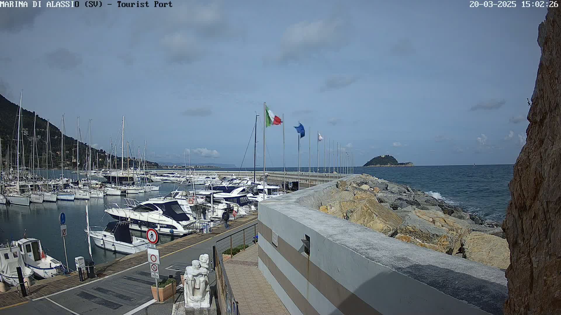 A marina filled with boats is situated next to a rocky shoreline, under a partly cloudy sky, with a calm sea beyond.