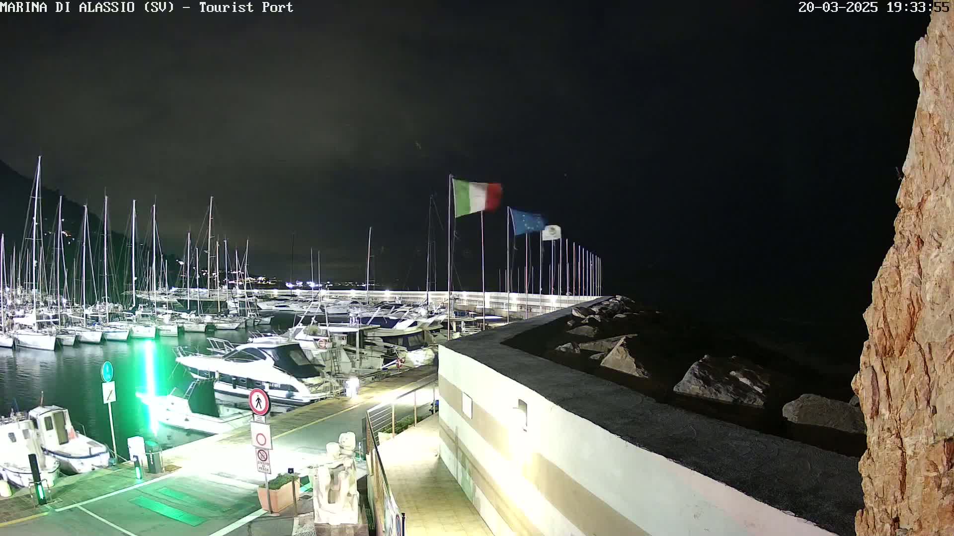 A nighttime view of a marina filled with boats, under a dark sky, with Italian and European flags waving on a nearby pier.