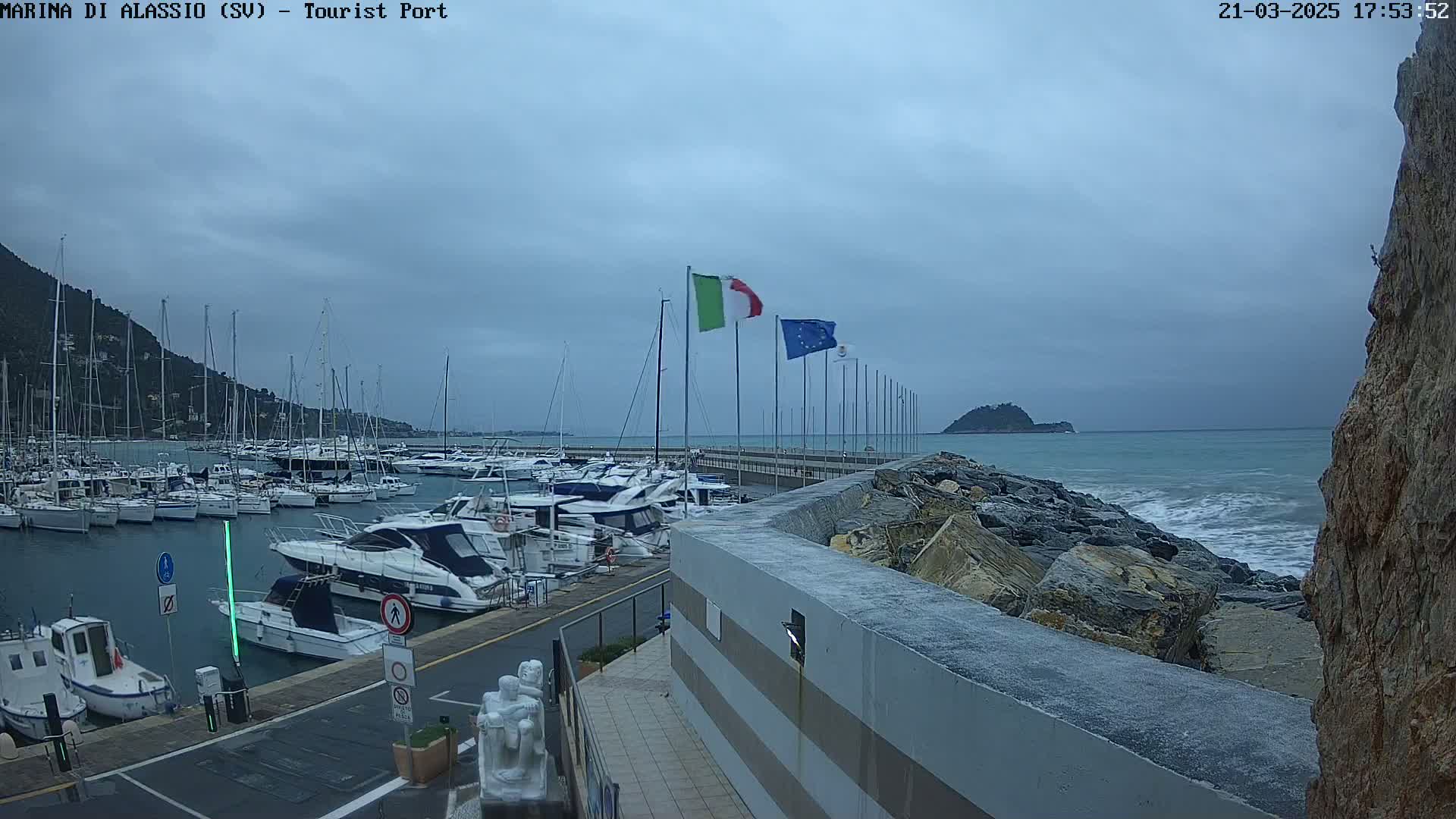 A marina filled with sailboats and motorboats is situated next to a rocky shoreline with small waves under a cloudy sky.