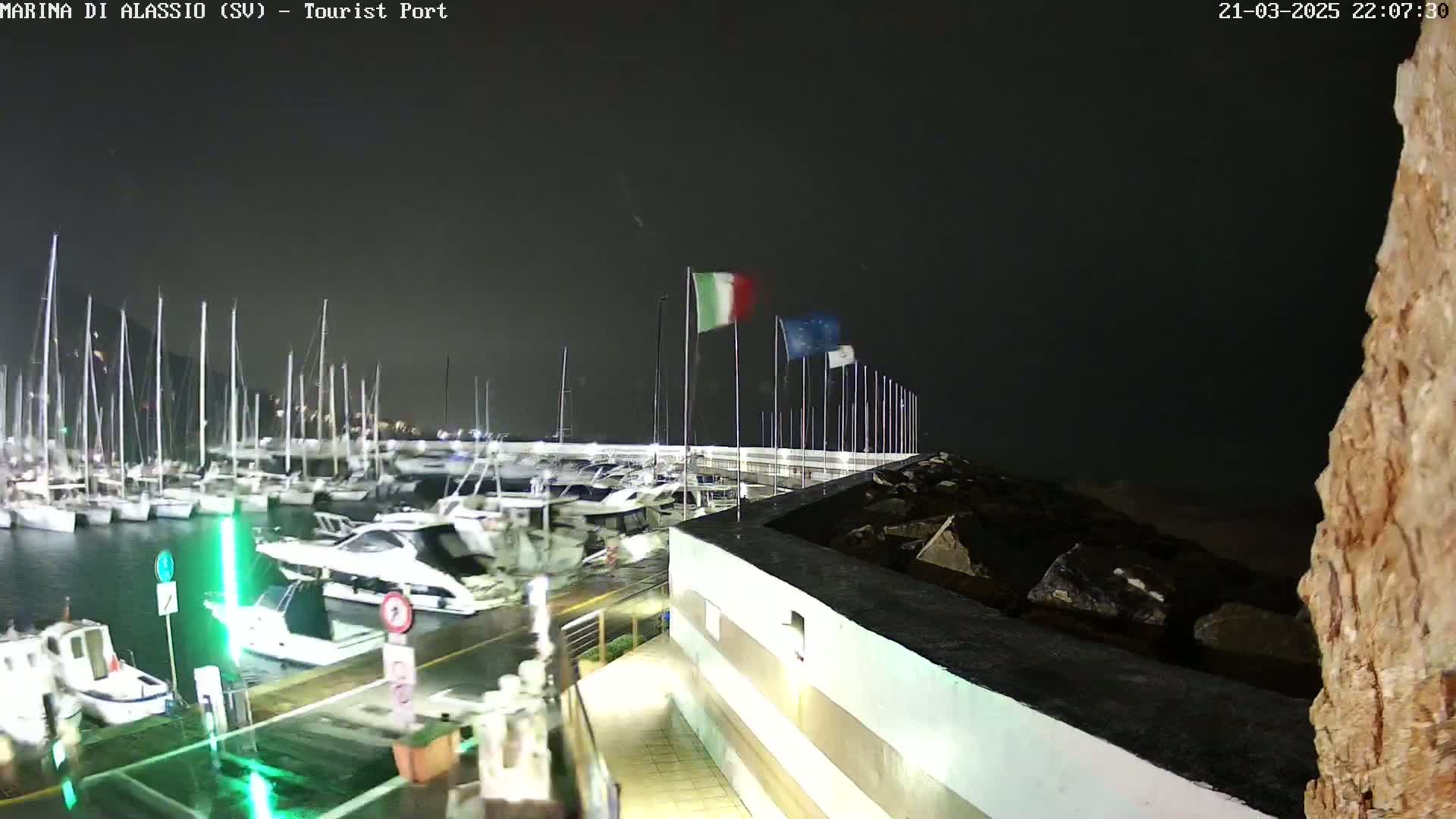 A nighttime view of a marina filled with boats, under a dark, possibly stormy sky, with flags waving in the wind.