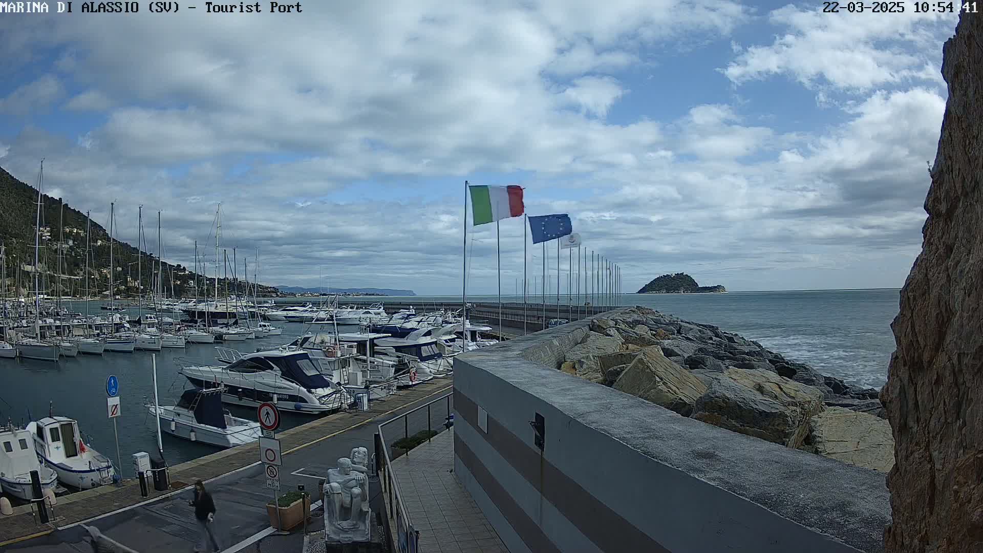 A marina filled with boats is situated on a partly cloudy day next to a rocky shoreline and a calm sea with a small island in the distance.