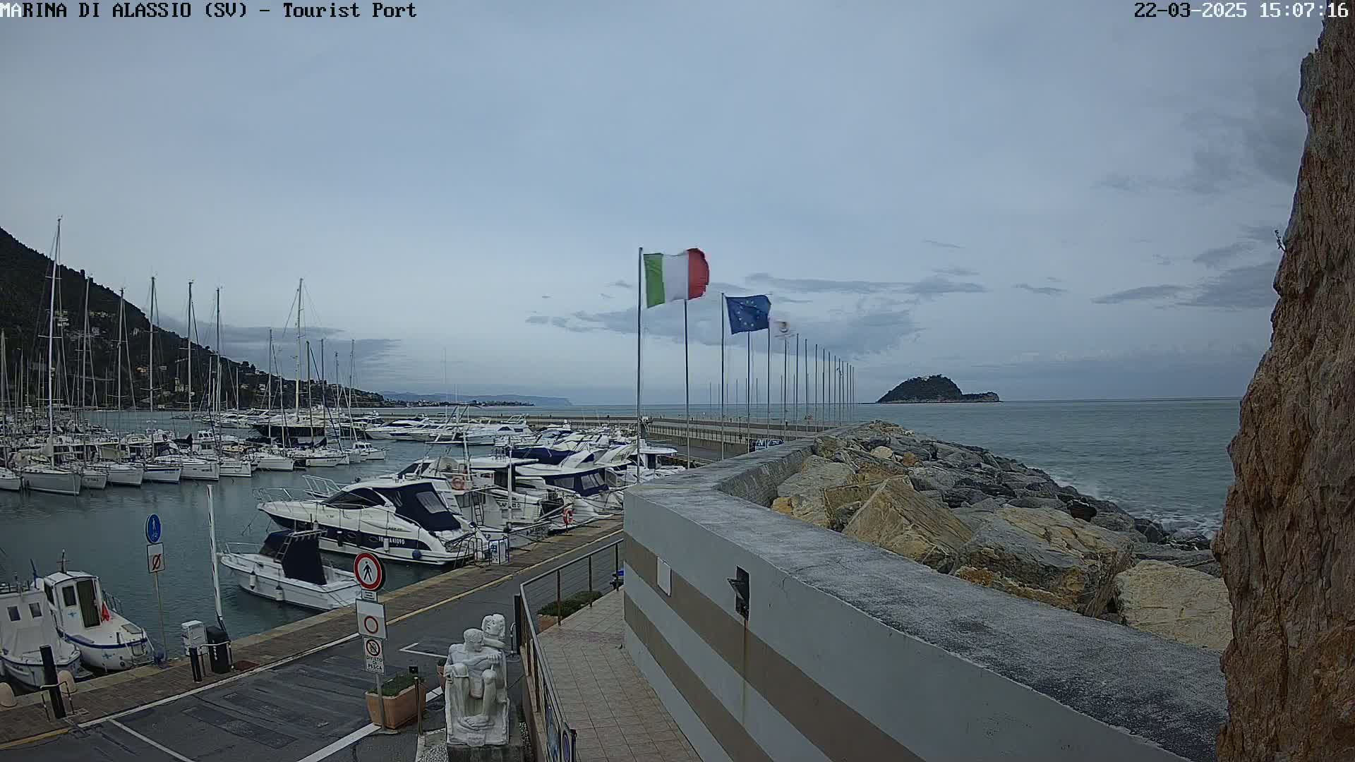 A marina filled with sailboats and motorboats is situated next to a rocky breakwater on a partly cloudy day.