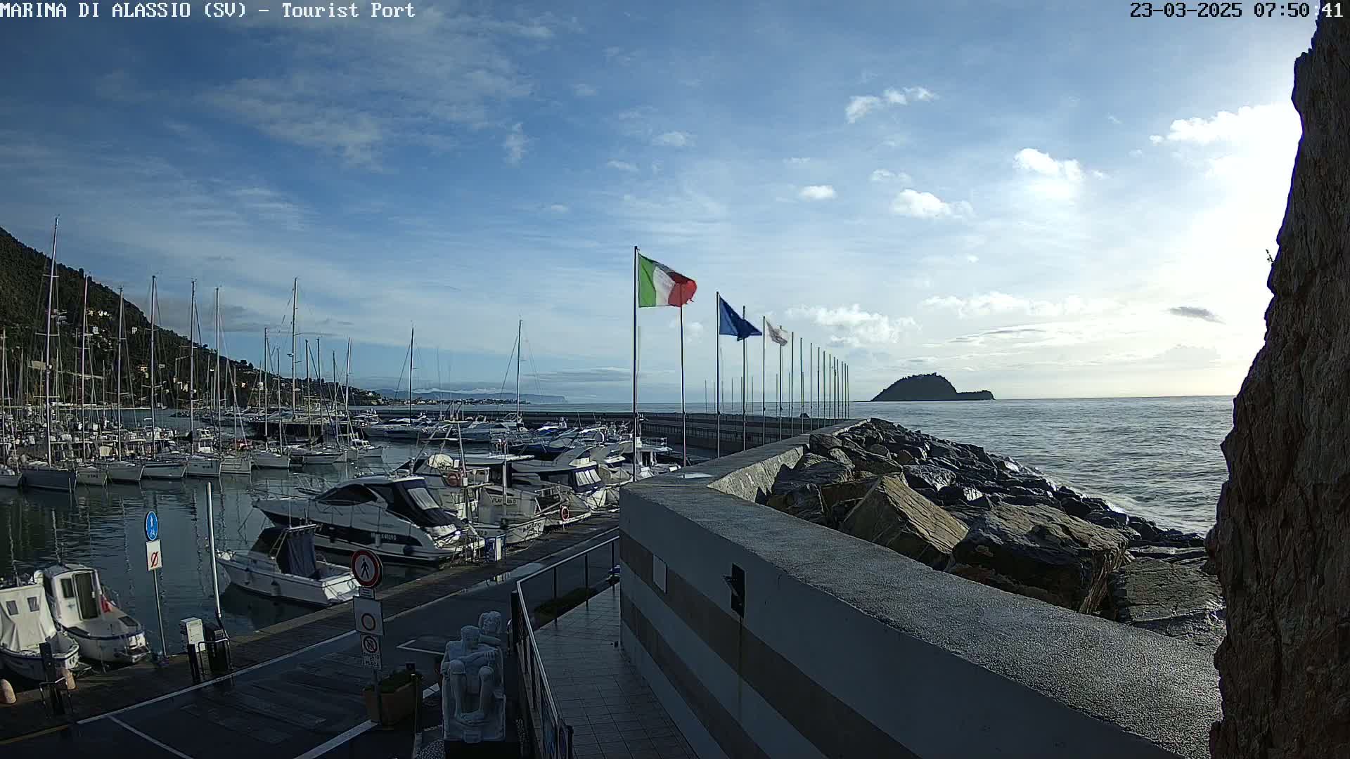 A marina filled with boats sits on a calm sea under a mostly sunny sky, with a rocky breakwater and a small island visible in the distance.