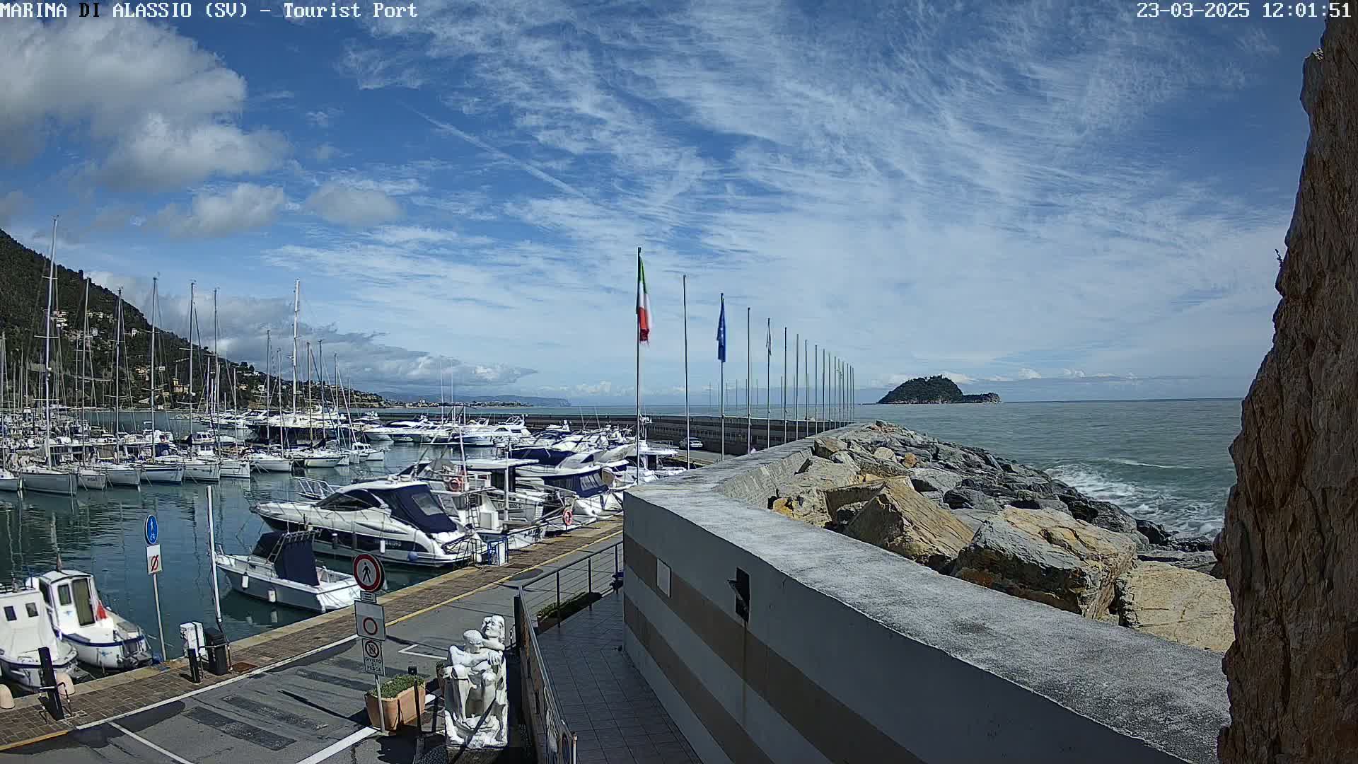 A marina filled with sailboats and motorboats is situated along a rocky coast under a partly cloudy sky, with gentle waves visible in the ocean.