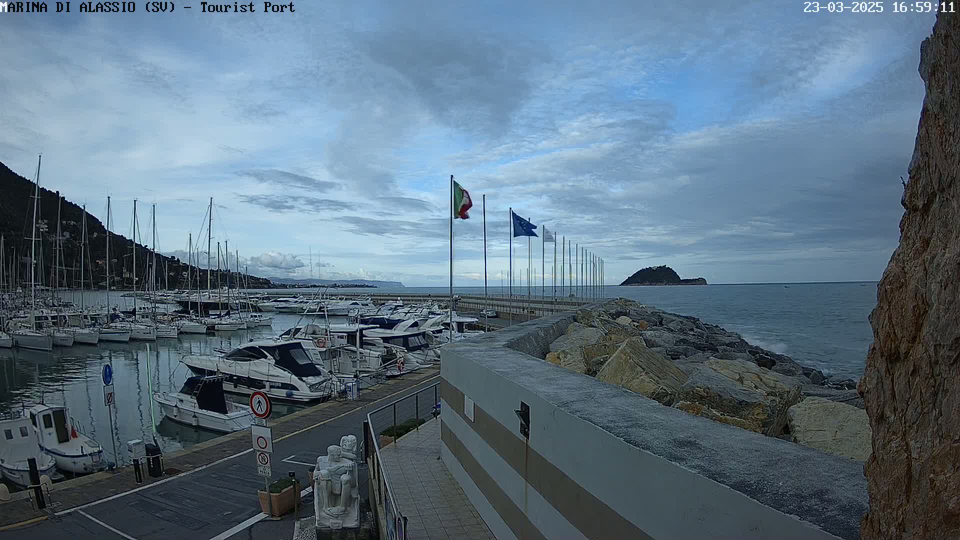 A marina filled with sailboats and motorboats is situated next to a rocky shoreline under a partly cloudy sky.