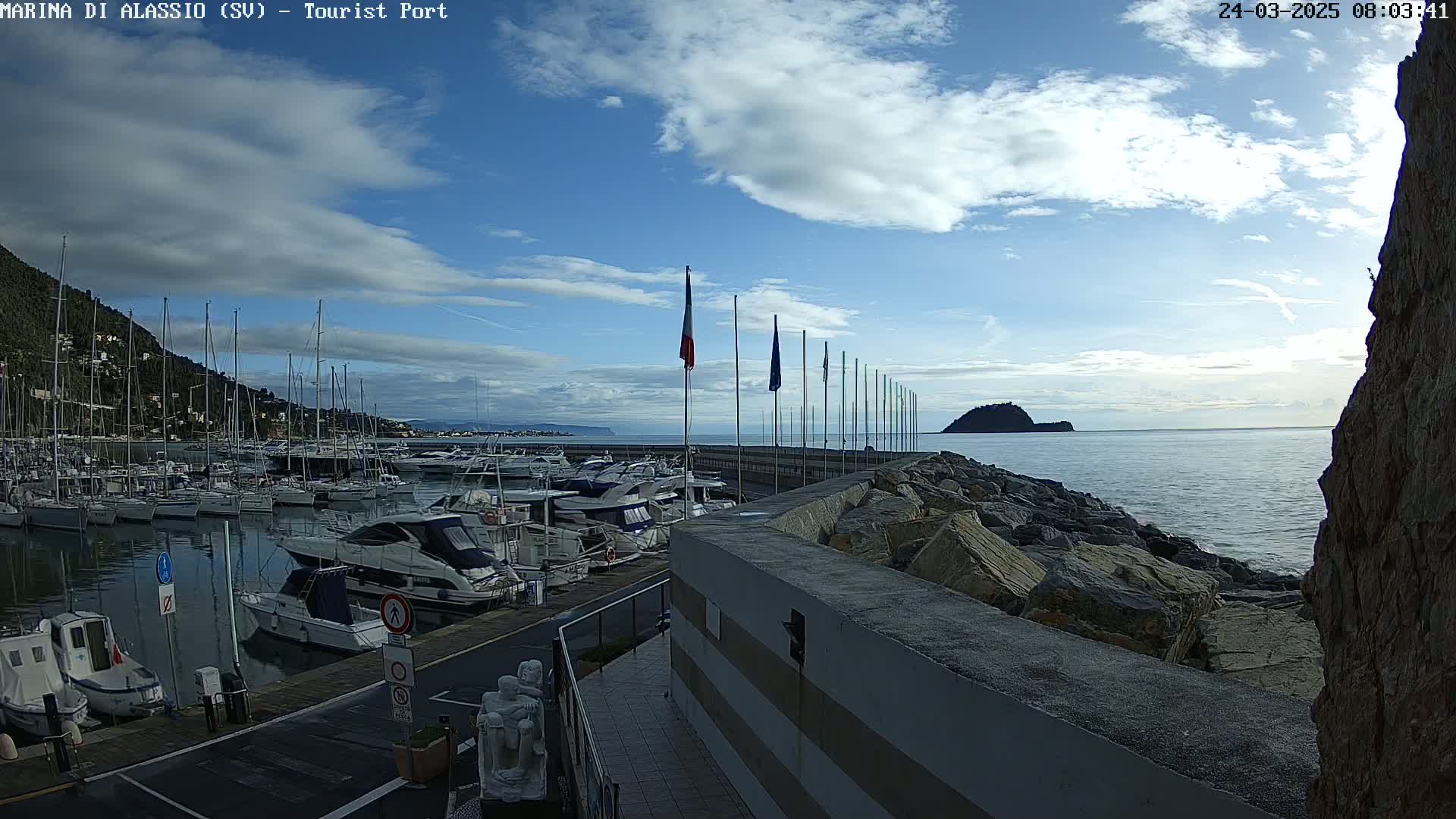 A marina filled with sailboats and motorboats is situated on a calm sea under a partly cloudy sky.
