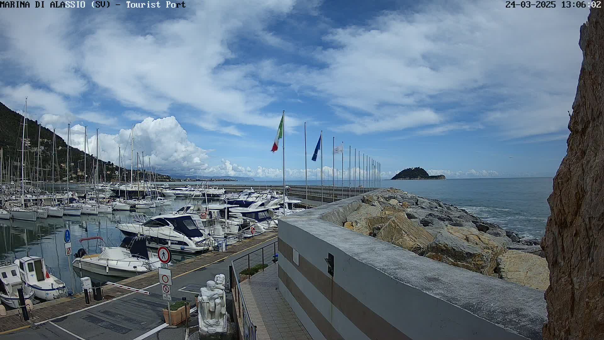 A marina filled with boats is situated next to a rocky shoreline under a partly cloudy sky.