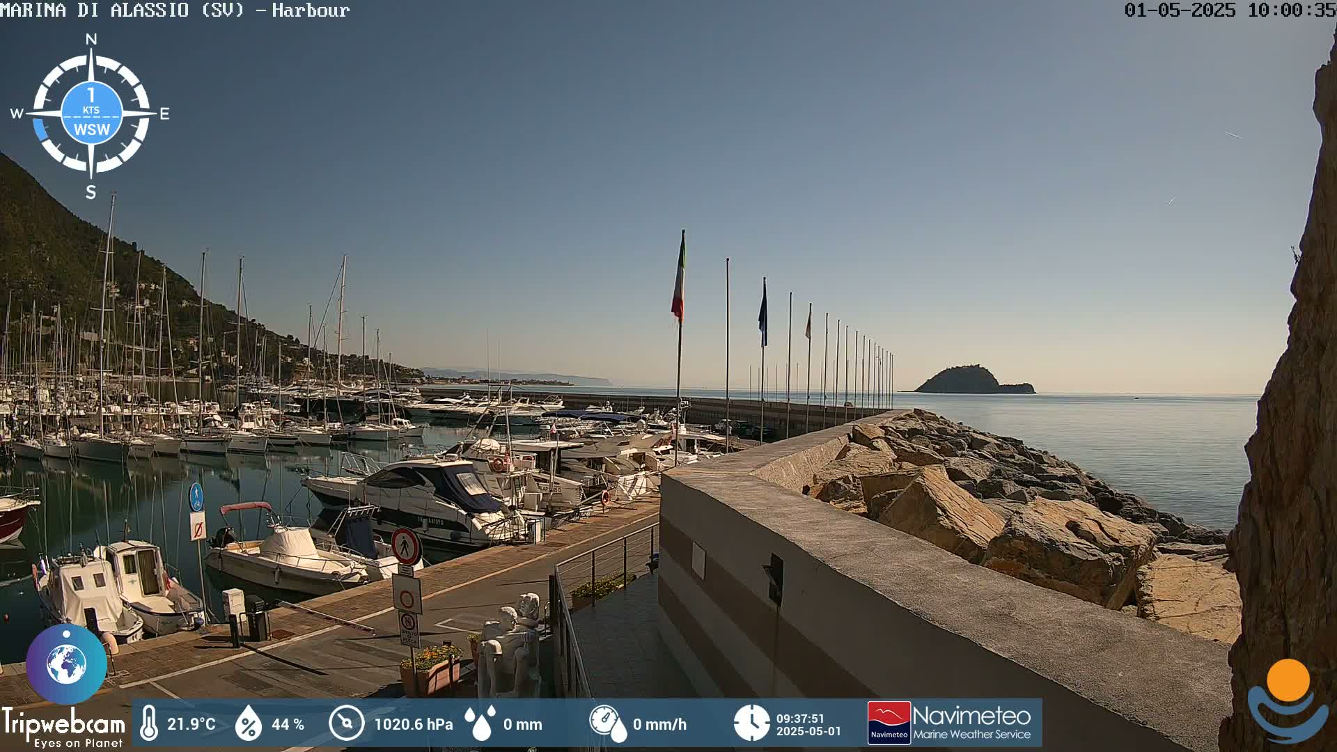 A harbor filled with numerous boats is situated next to a rocky shoreline, under a clear blue sky.
