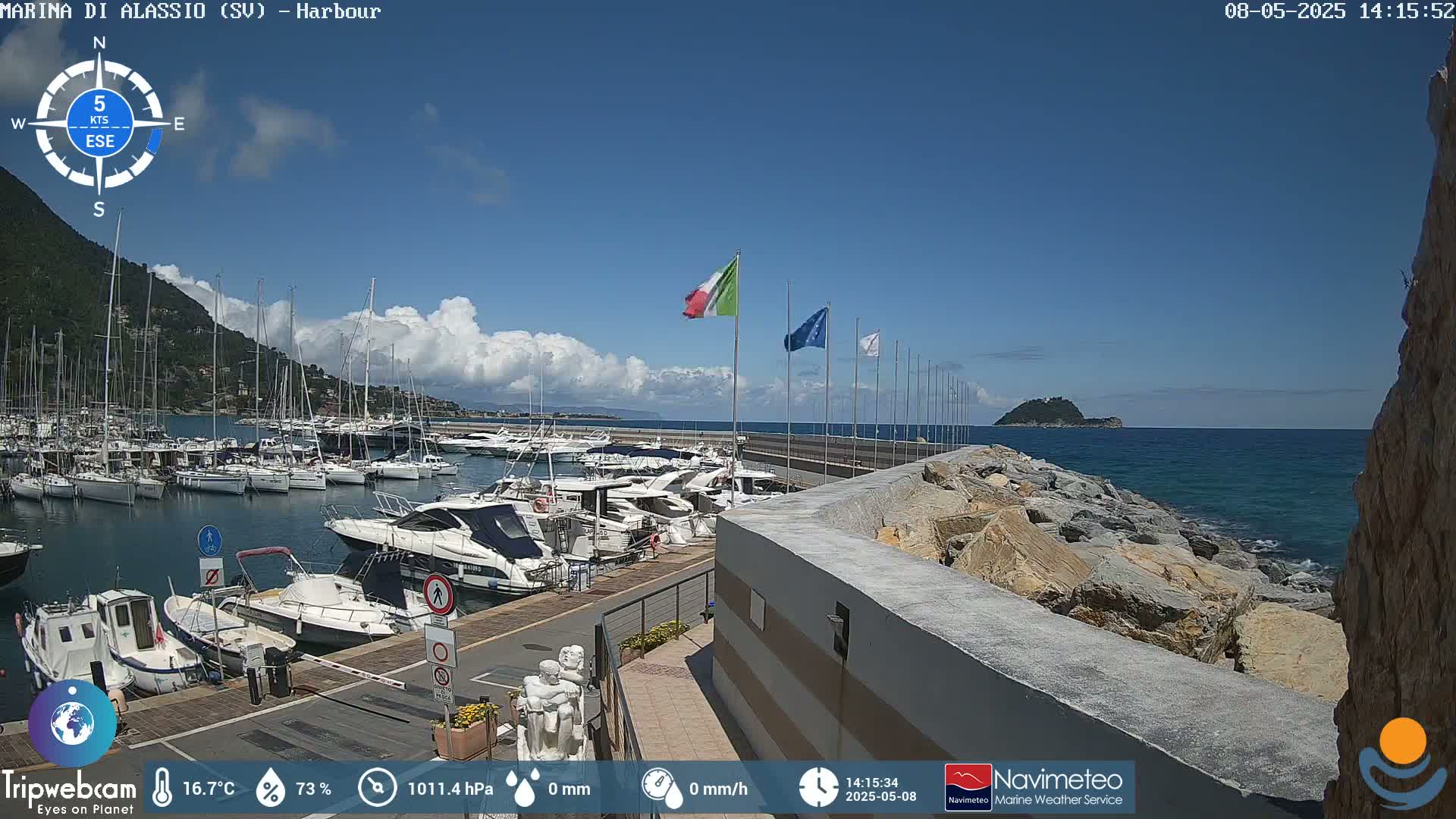 A harbor filled with sailboats and motorboats is situated near a rocky shoreline under a mostly sunny sky with some scattered clouds.