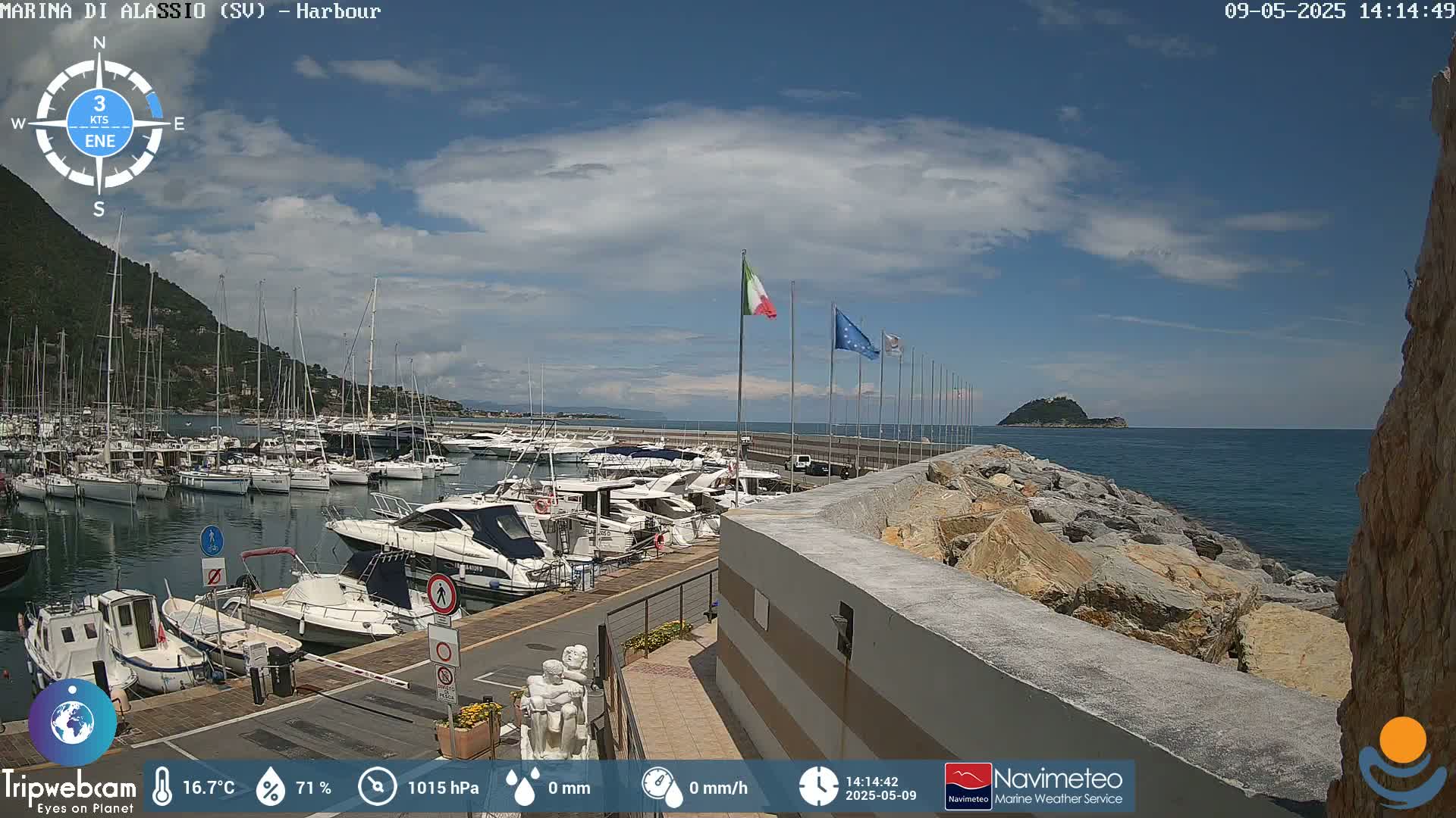 A harbor filled with numerous boats is situated on a calm, sunny day next to a rocky shoreline and a small island in the distance, under a partly cloudy sky.