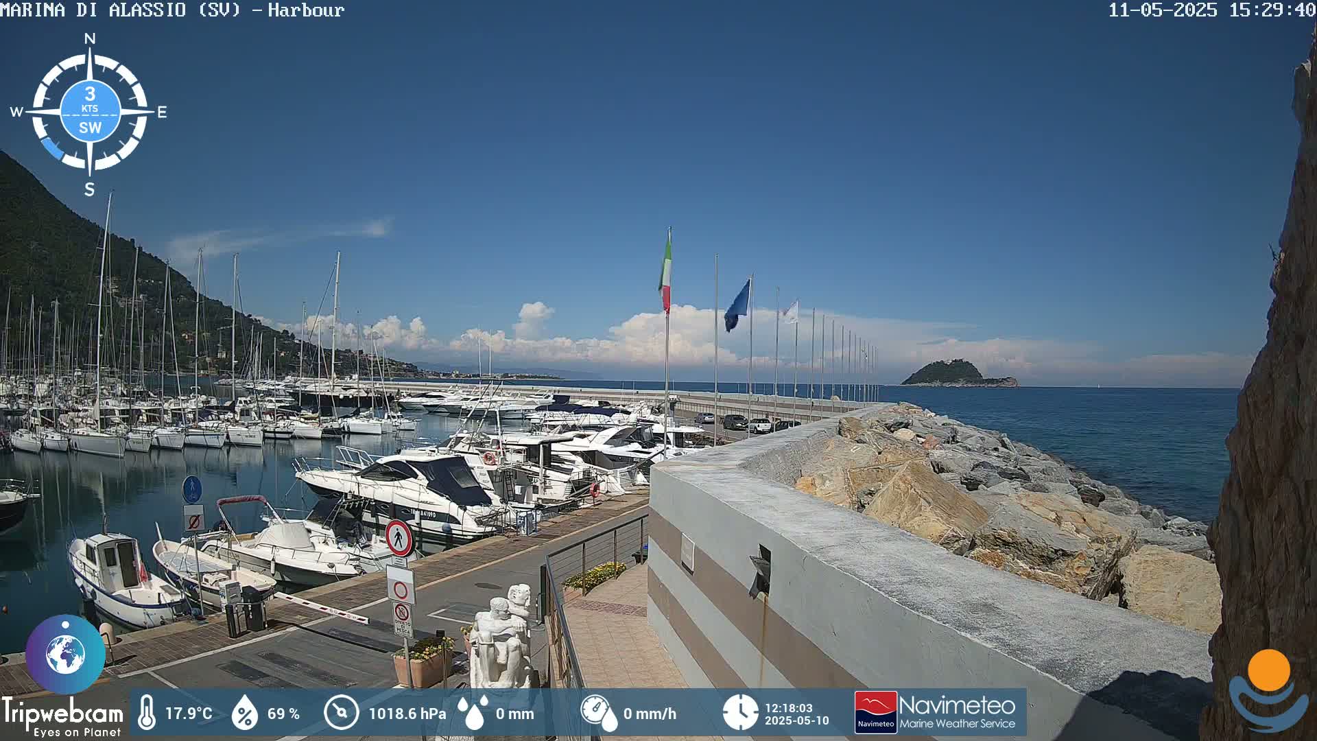 A harbor filled with boats is seen on a sunny day with some clouds, backed by a hill and an island in the distance.