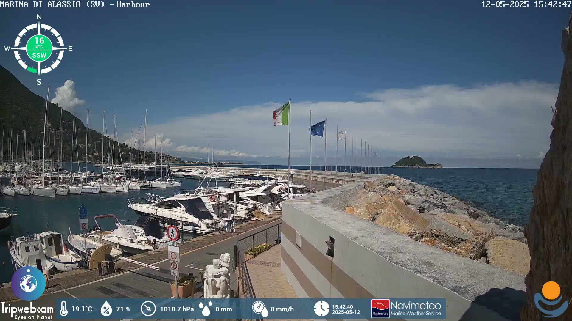 A harbor filled with boats is situated under a mostly sunny sky with a few clouds, next to a rocky shoreline and a small island in the distance.