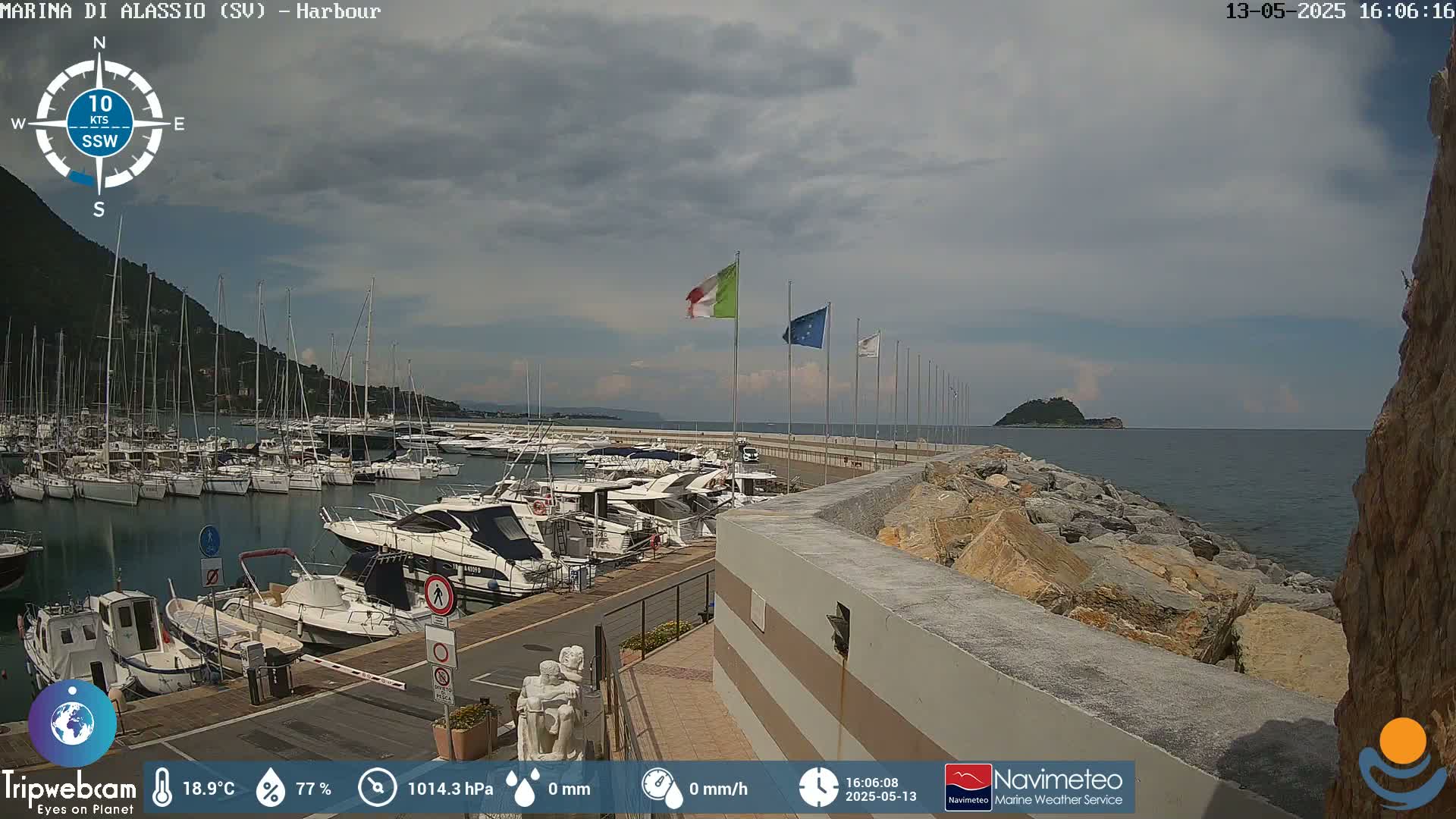 A partly cloudy day shows a harbor filled with numerous boats, a rocky shoreline, and a small island in the distance.