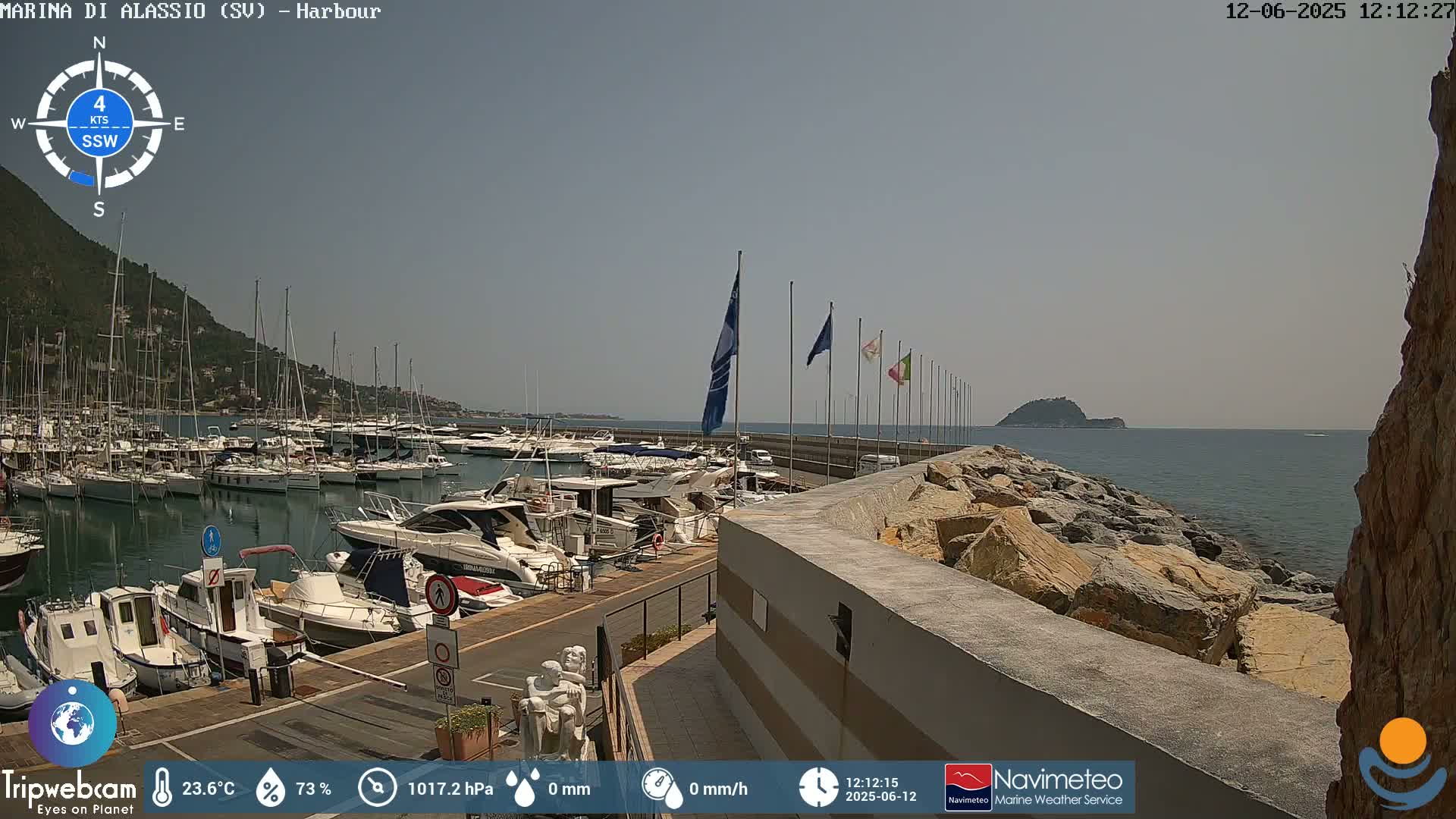 A harbor filled with numerous boats is situated next to a rocky shoreline on a sunny day.