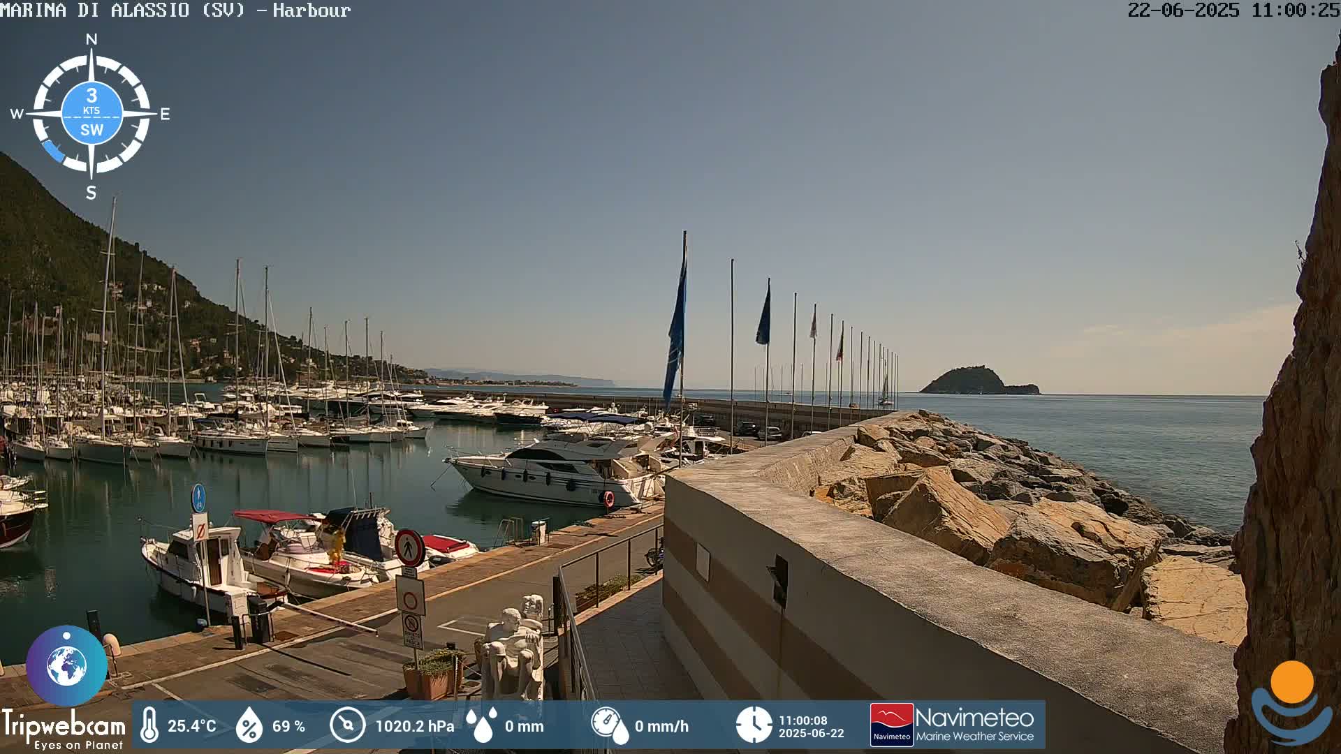 A harbor filled with numerous boats and yachts is shown under a sunny sky, with a rocky breakwater extending into the calm sea.