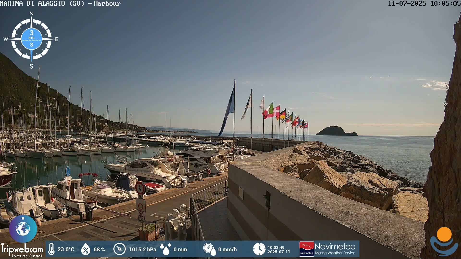 A marina filled with boats is situated next to a rocky seawall with numerous flags, under a clear sky on a sunny day.