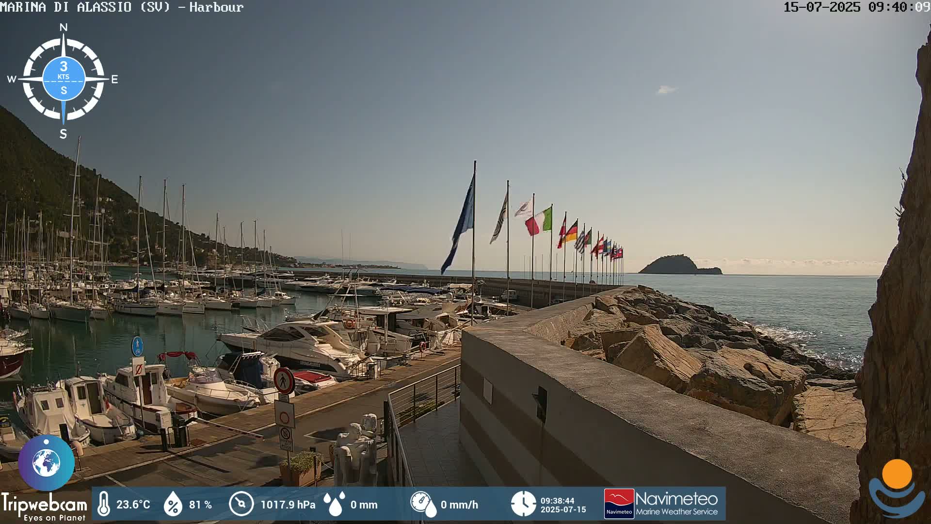 A harbor filled with boats is situated on a calm, sunny day next to a rocky shore with a small island in the distance, under a clear sky.