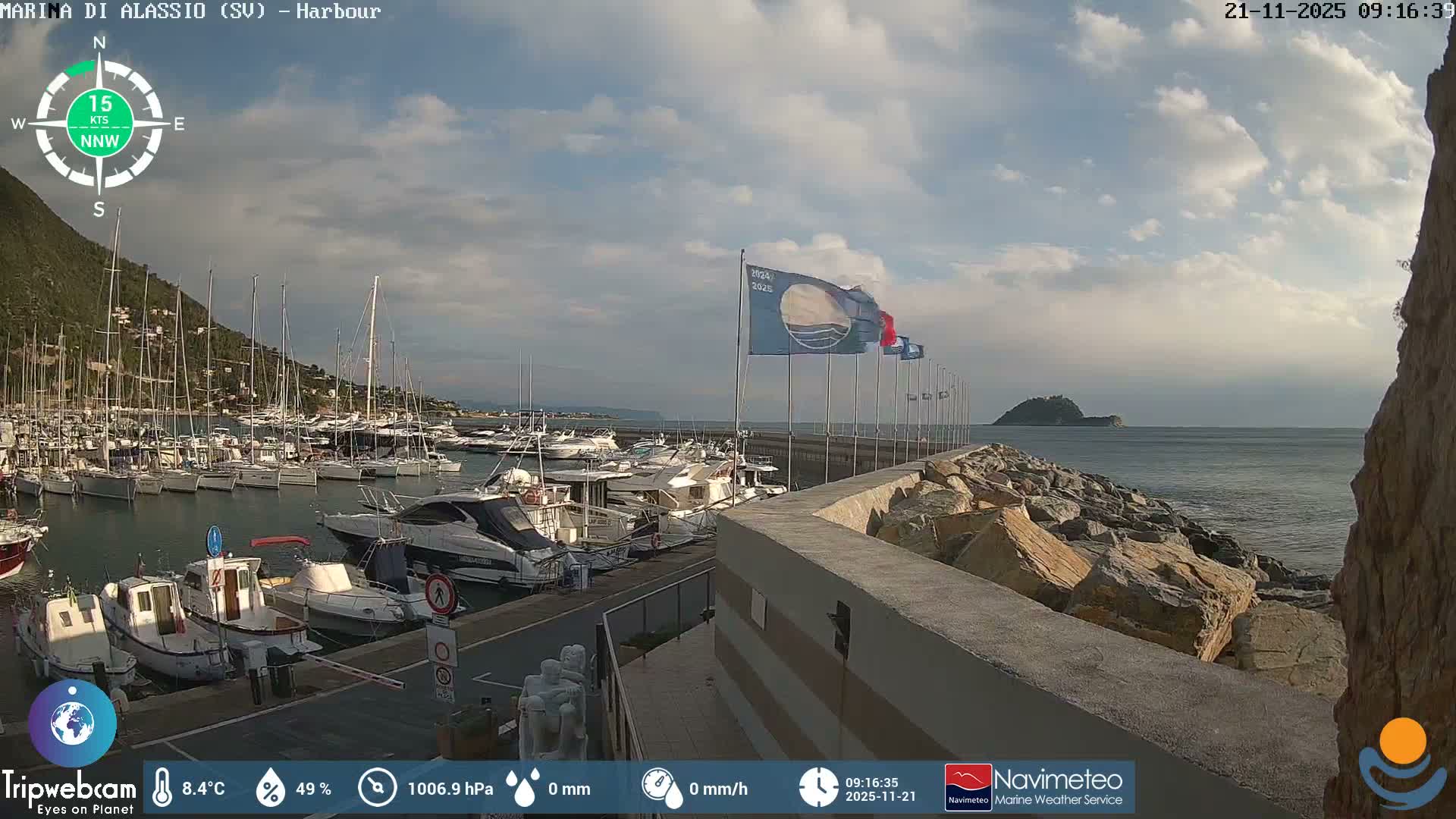 A harbor filled with numerous boats is visible under a partially cloudy sky, with a green hillside to the left, a rocky breakwater extending into a calm sea, and a small island in the distance, indicating a mild, breezy day.