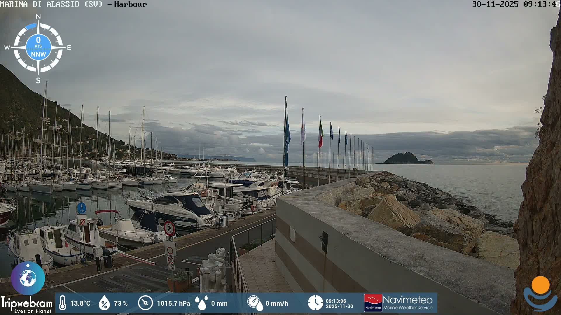 A wide view of a crowded marina on an overcast day shows numerous boats docked in calm waters, with a steep green mountain rising on the left, a pier lined with flags, a rocky breakwater, and a distant island in the cloudy sea.