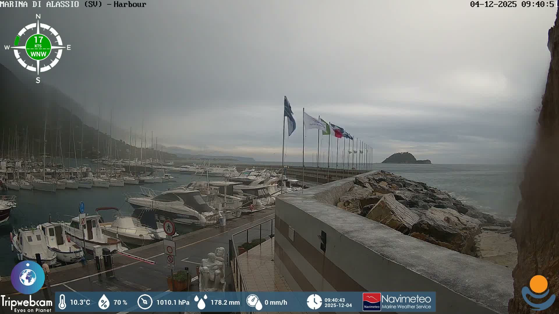 Under a heavy, overcast sky, a marina is filled with numerous boats and yachts alongside a wet pier and rocky breakwater, with distant hills and an island visible across choppy waters.