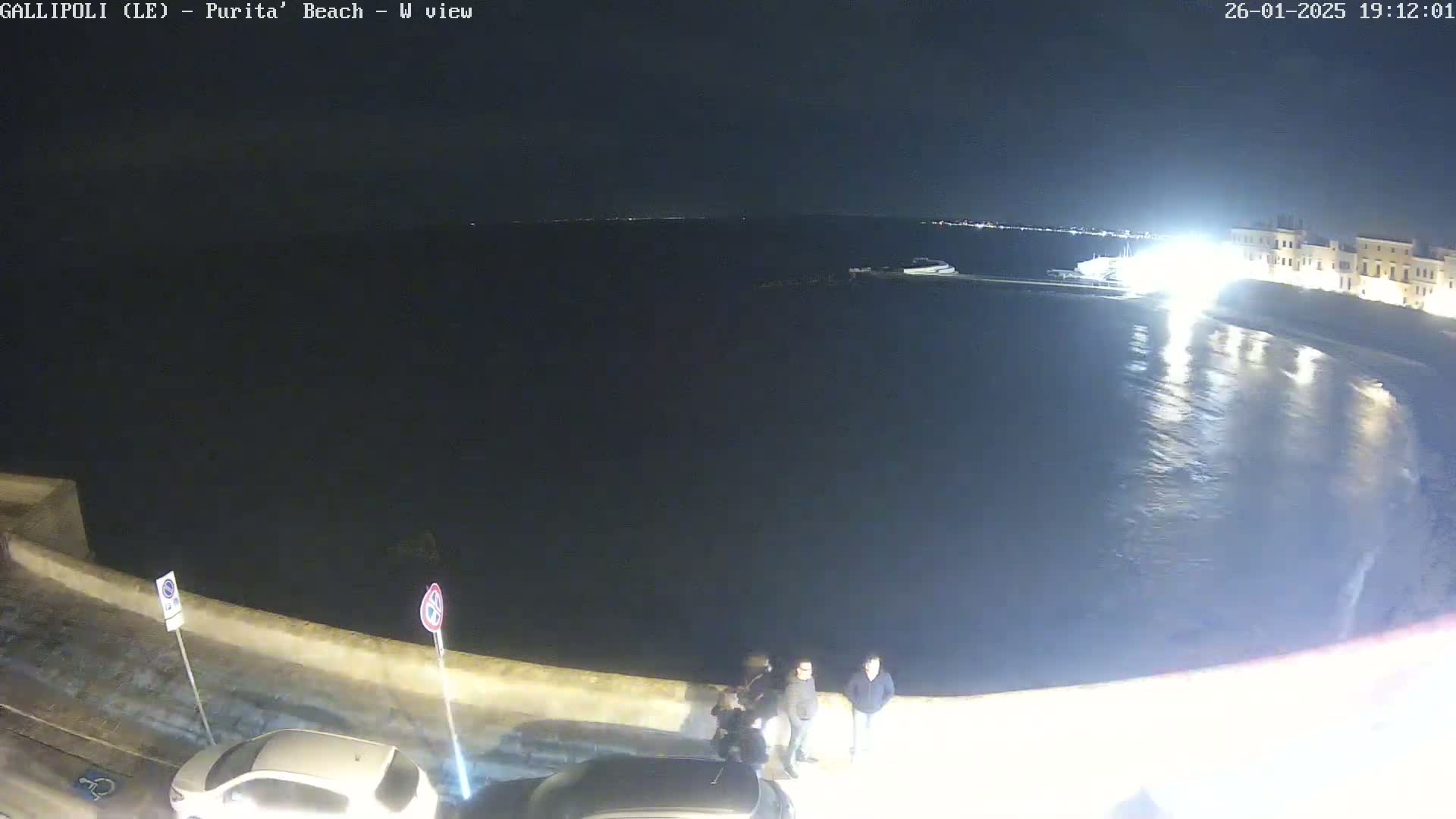 A nighttime view of a calm, dark ocean next to a stone wall with several people standing near parked cars.