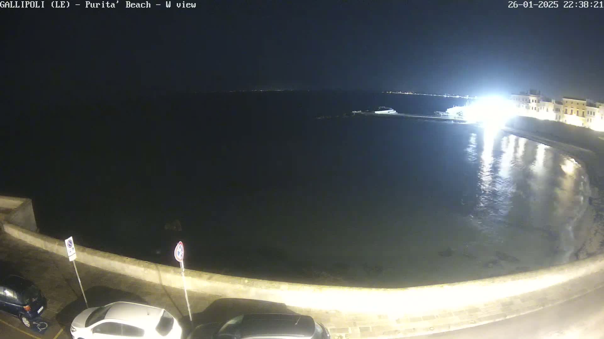 A nighttime view of a calm body of water next to a stone wall and parked cars under a clear, dark sky.