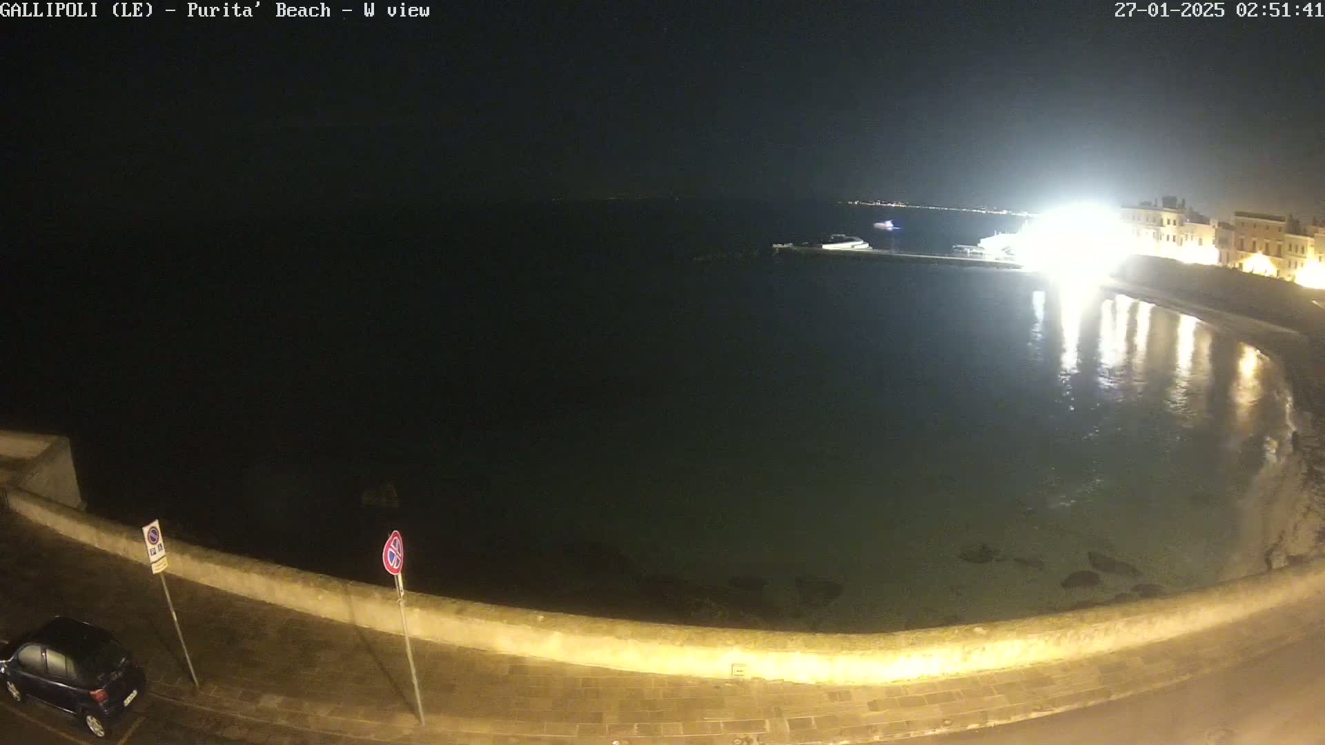 A nighttime view of a calm, dark body of water next to a low stone wall, with a few lights visible in the distance and a car parked on the road.