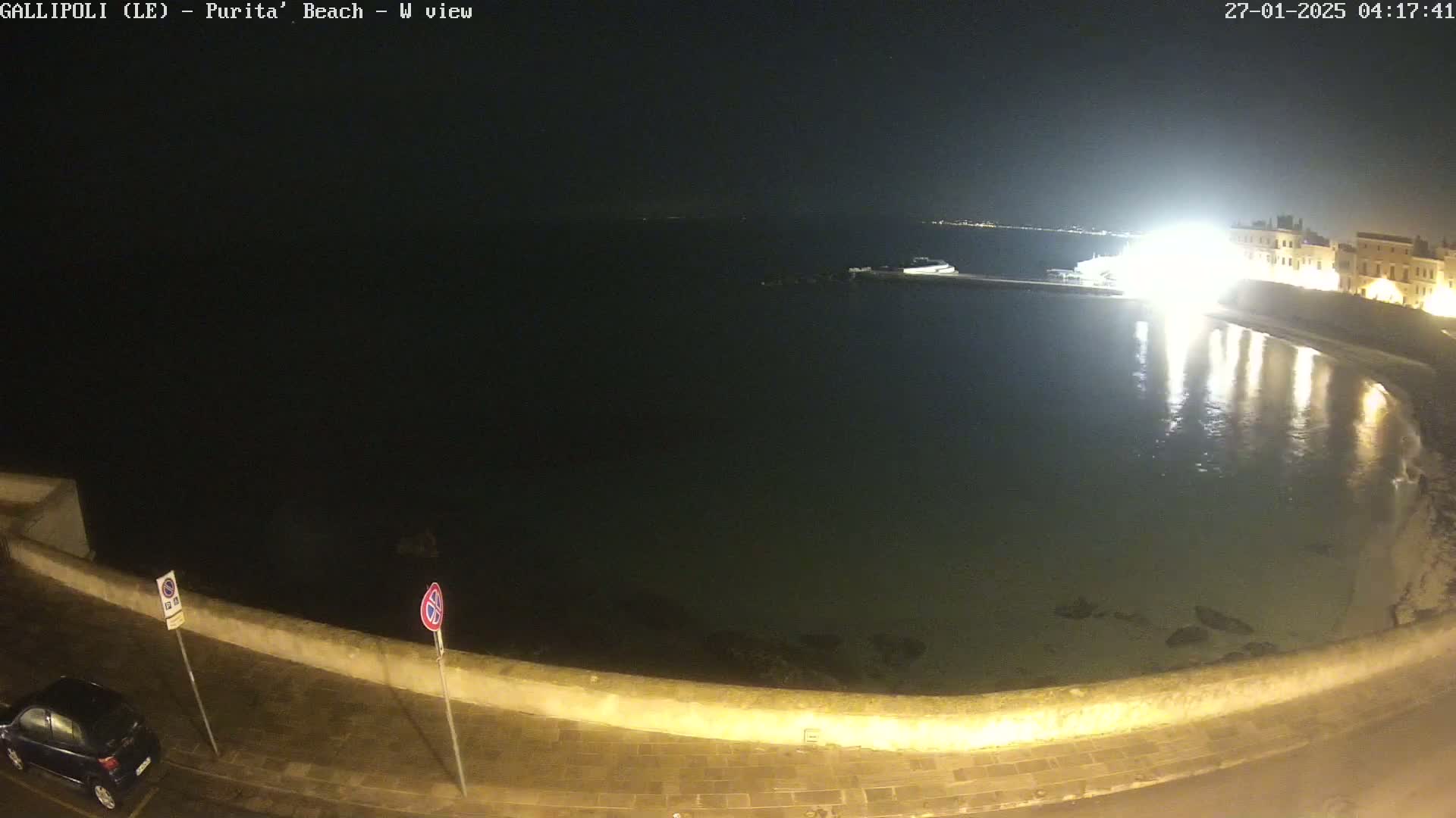 At night, a dark body of water is seen next to a low wall, with buildings and lights visible in the distance.