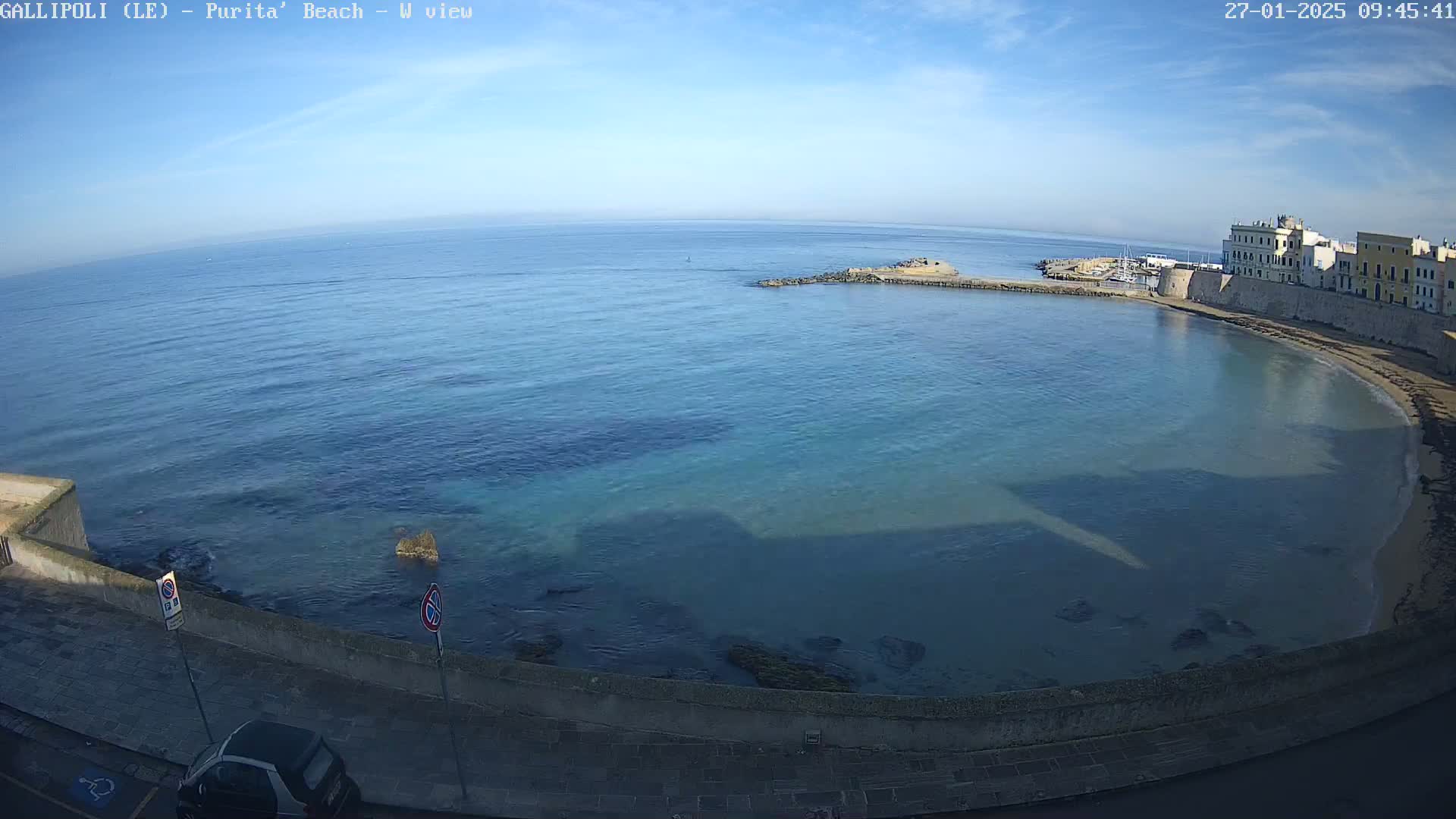 A high-angle view shows a calm, clear blue ocean curving into a small sandy beach next to a stone wall and buildings under a sunny sky.