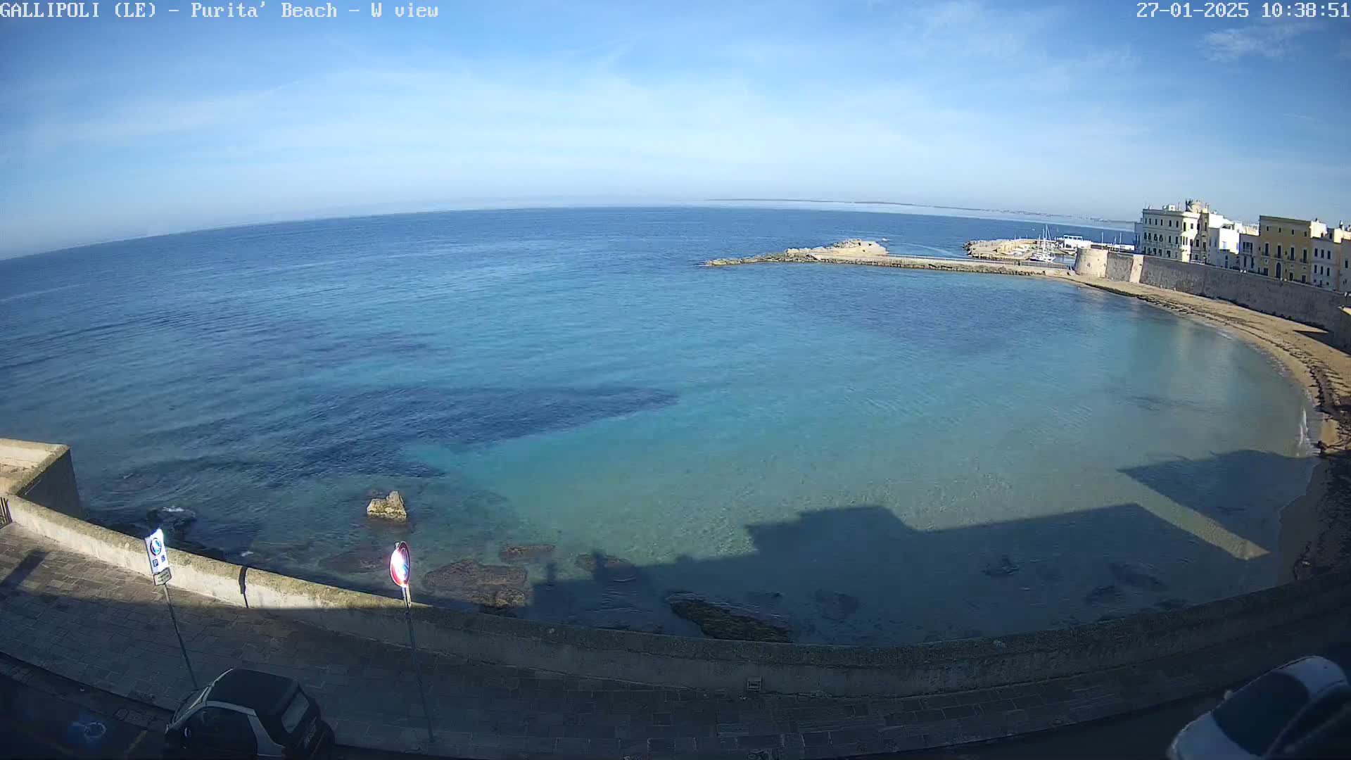 A clear, sunny day shows a calm, turquoise bay curving around a stone city wall, with cars parked along the waterfront.