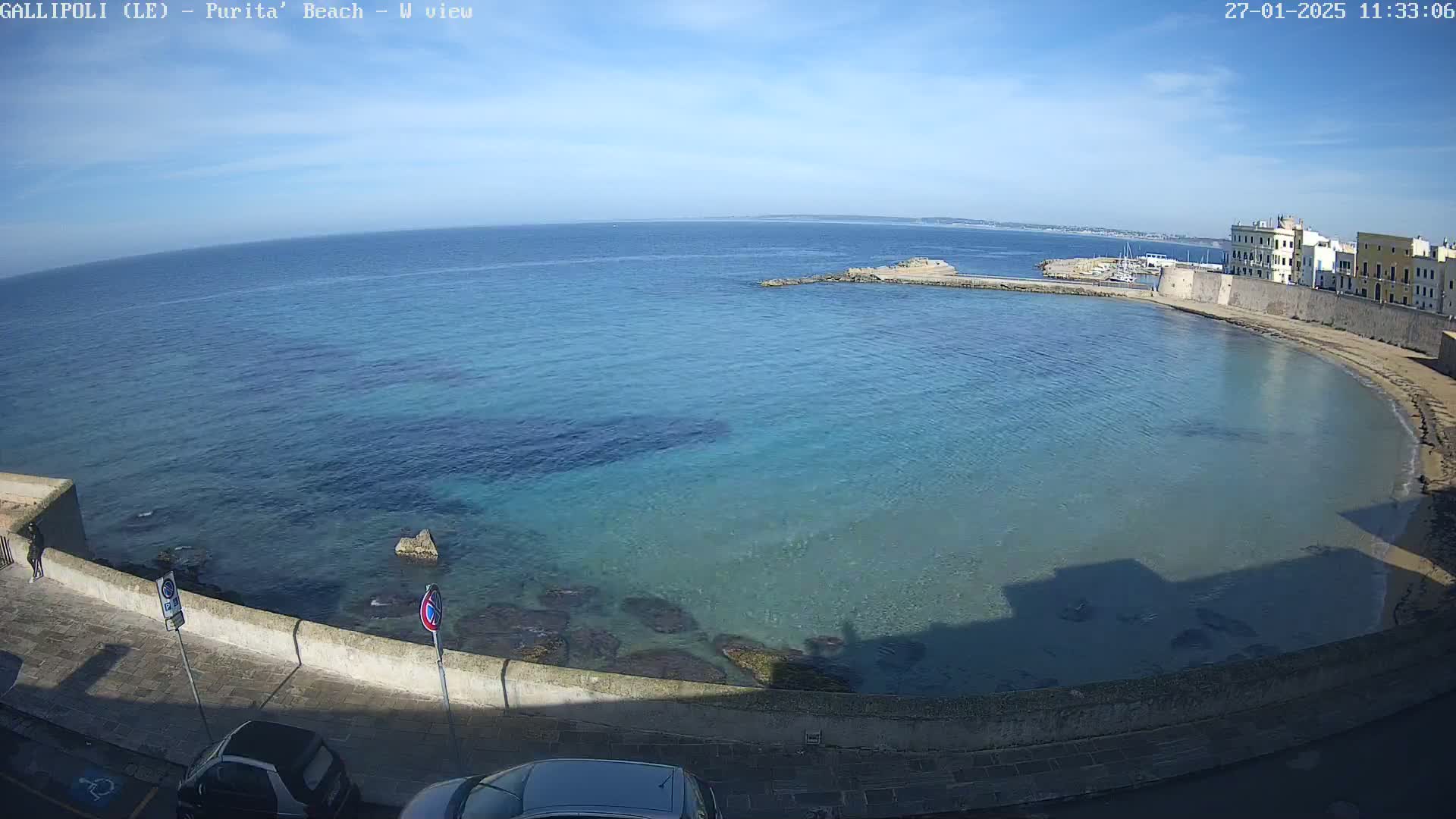 A clear, sunny day reveals a tranquil bay with clear, shallow turquoise water lapping a sandy shore next to a stone wall and buildings.