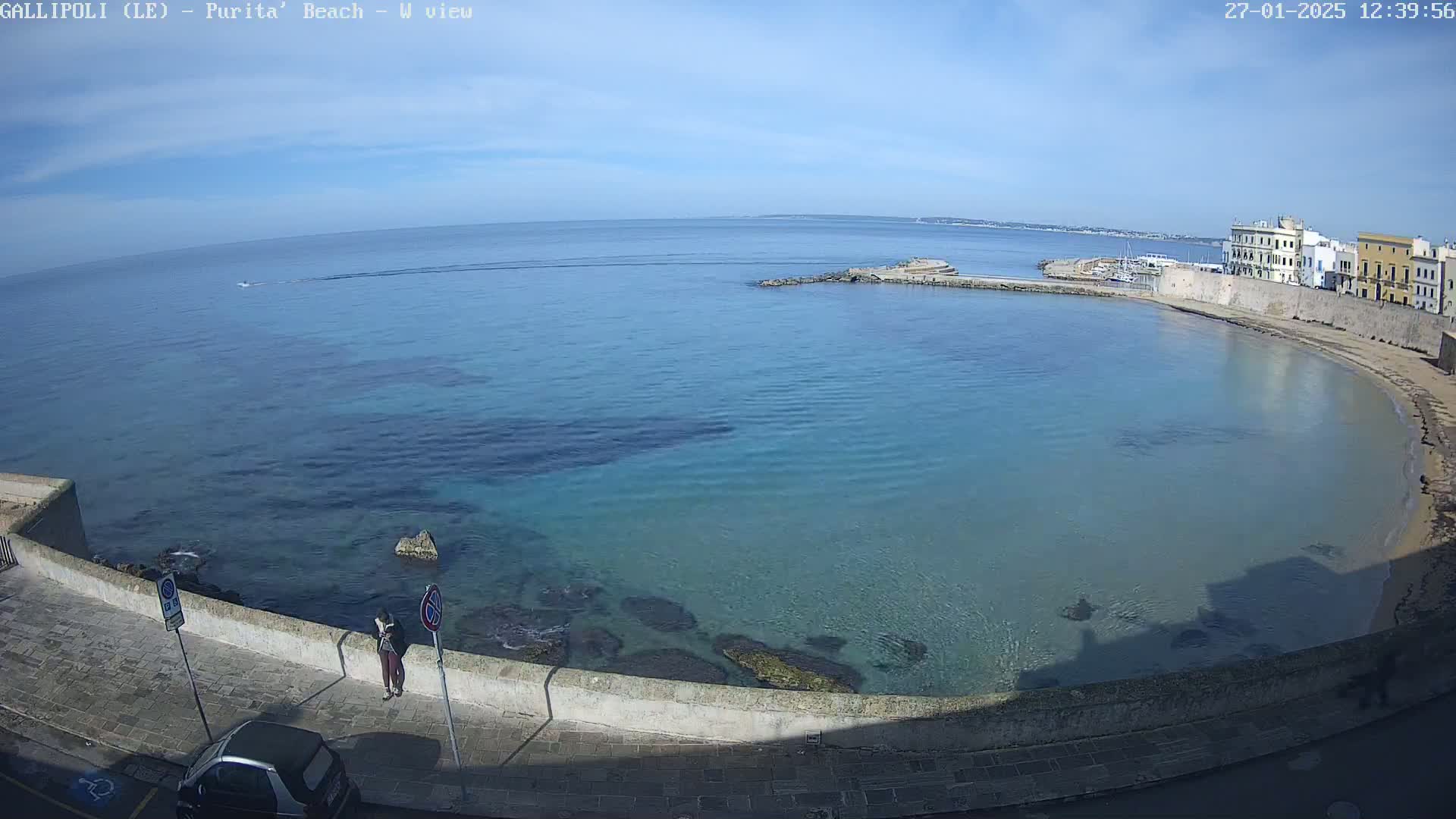 A clear, sunny day overlooks a calm, blue bay with a sandy beach and a stone wall bordering a paved walkway.