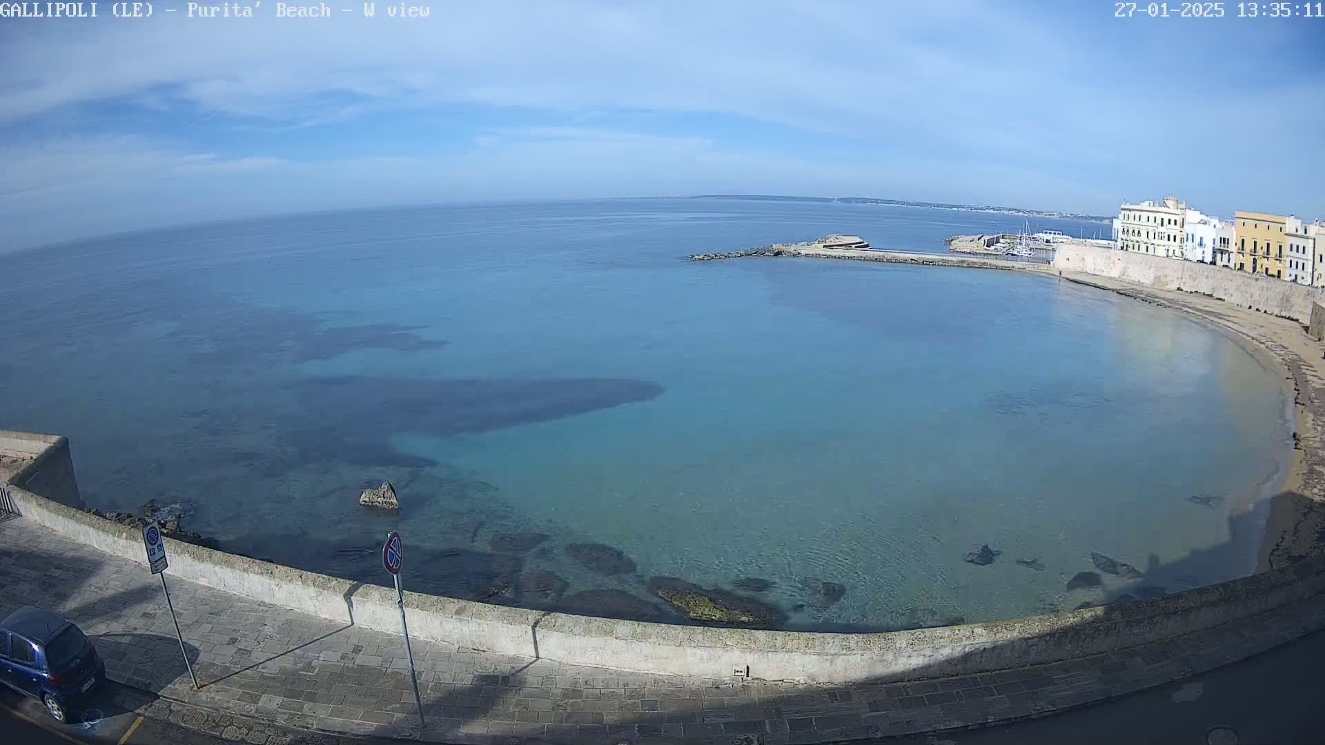 A clear, sunny day overlooks a calm, shallow bay with clear turquoise water, a stone wall, and buildings lining the shore.