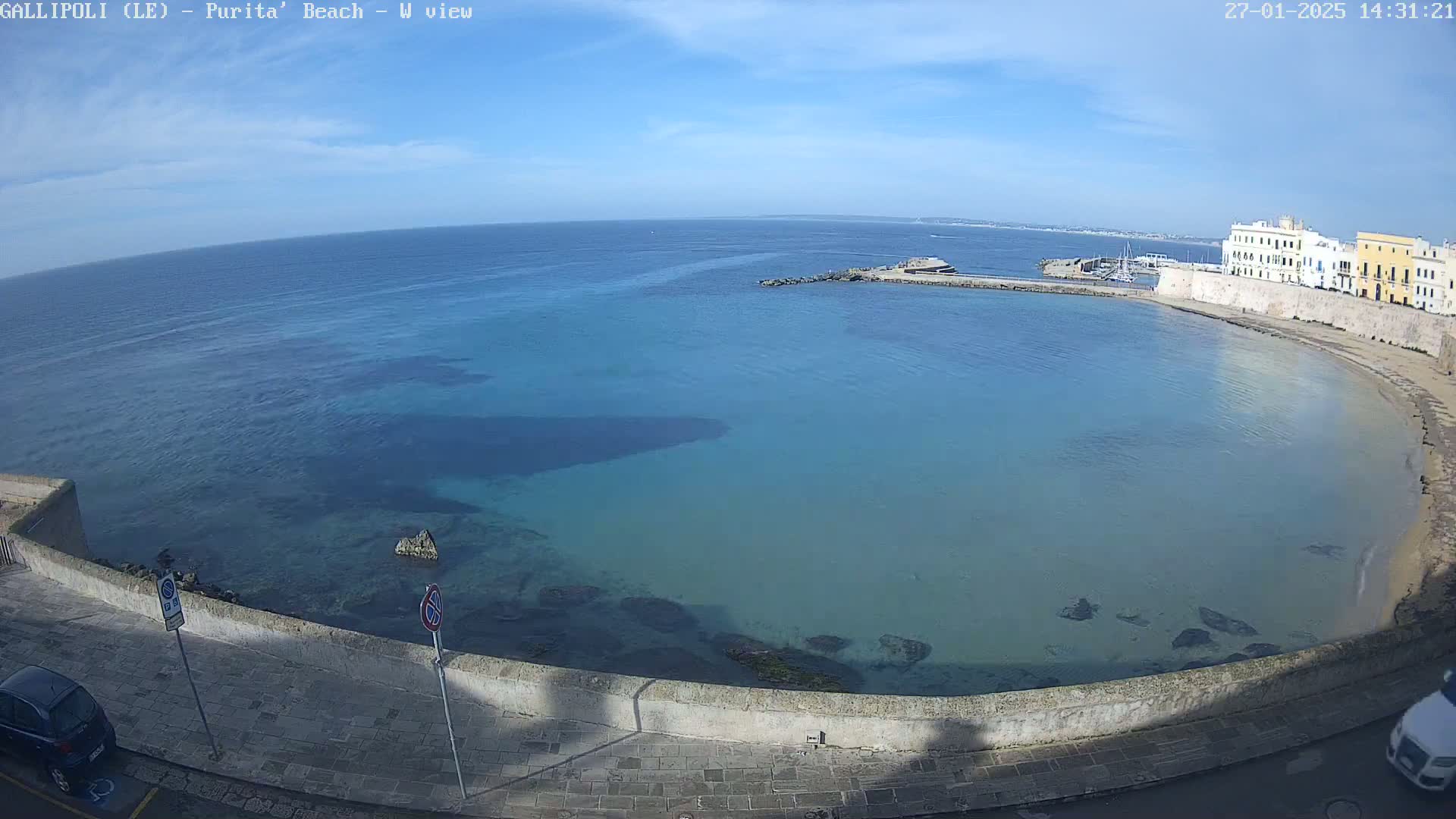 A clear, sunny day reveals a tranquil bay with pale turquoise water lapping a sandy beach beside a stone wall and buildings.