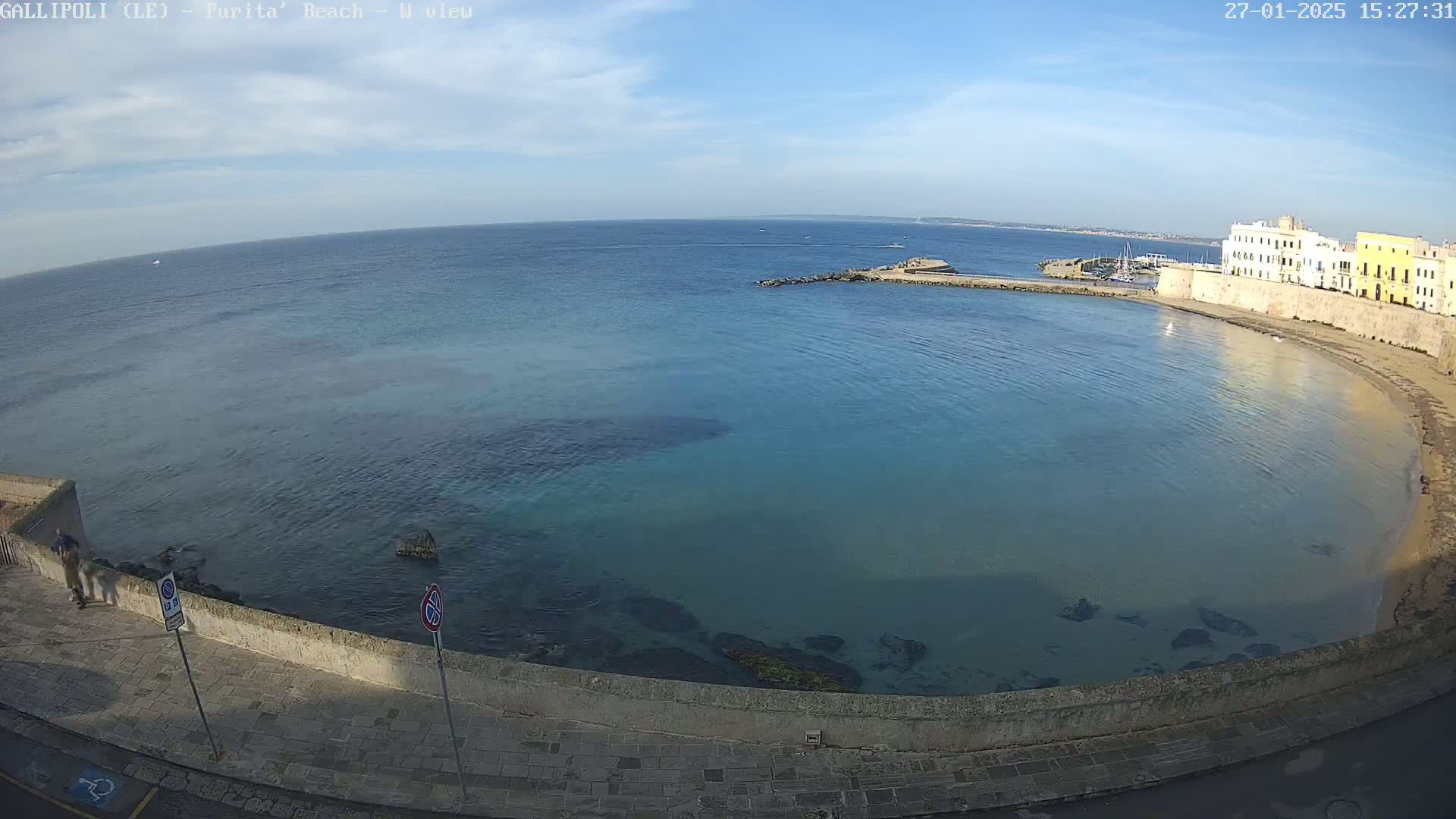 A clear, sunny day overlooks a calm, blue bay with a sandy beach and stone buildings along the shoreline.
