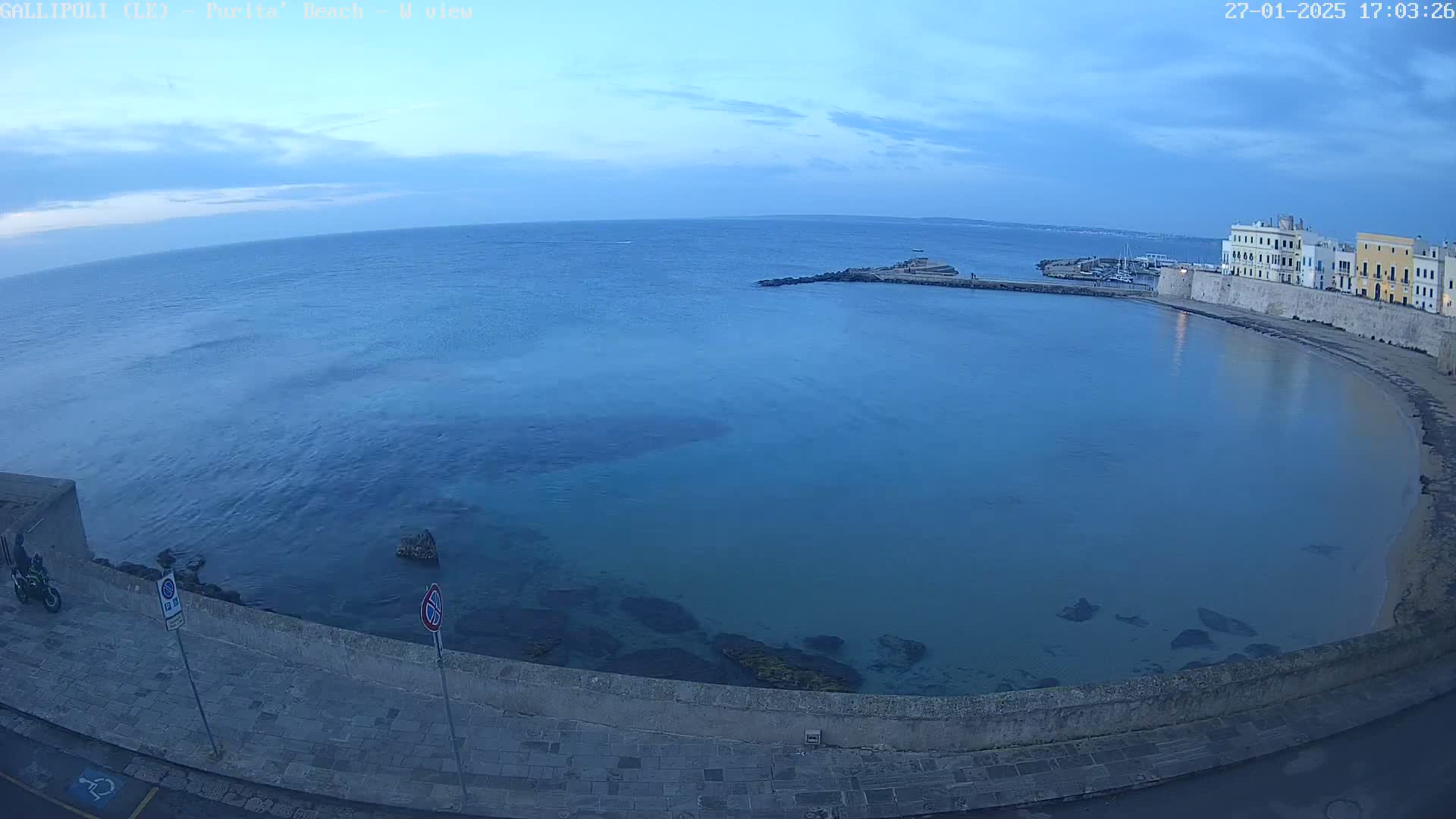 A calm, blue sea curves around a stone wall and beach at twilight, with a small town visible in the distance.