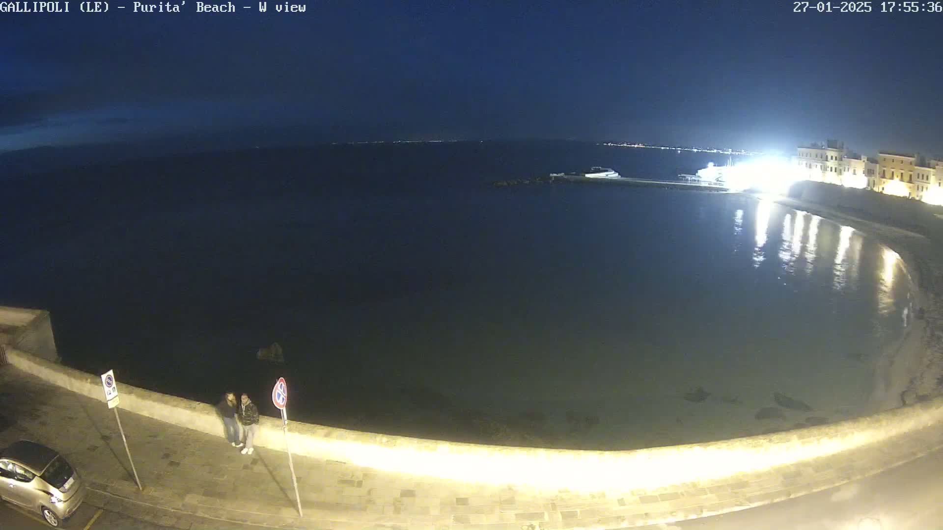 A nighttime view of a calm, dark body of water next to a low wall, with two people standing on the wall and a car parked nearby under a clear, dark sky.
